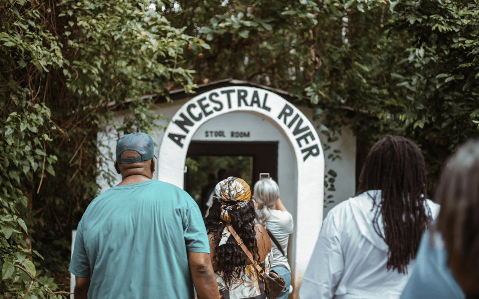 A group of people entering a building labeled 'Ancestral River' with a sign 'Stool Room' at the entrance, surrounded by green foliage in Ghana