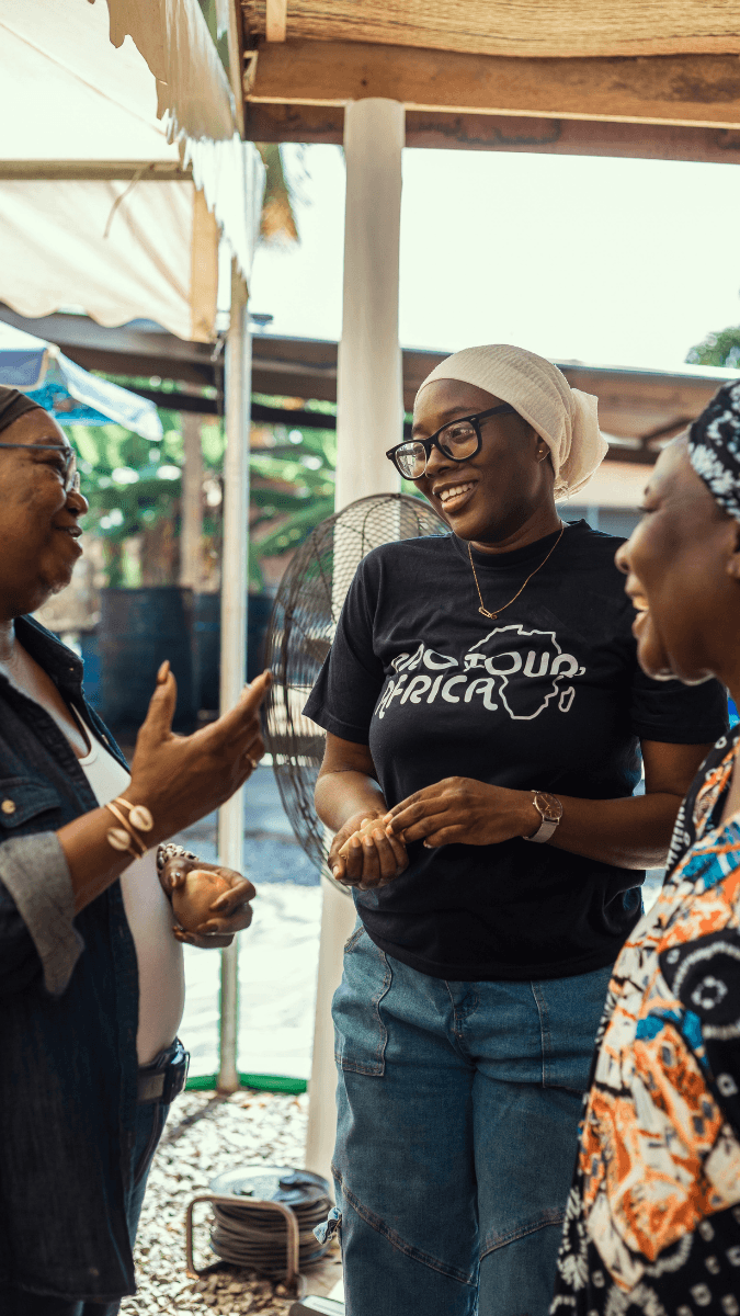 Three women engaged in conversation outdoors under a canopy, smiling and laughing, with one woman wearing a black T-shirt with Protour Africa logo