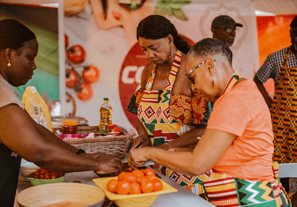 Three women at a cooking class in Ghana preparing tomatoes, surrounded by various food products and bottles.