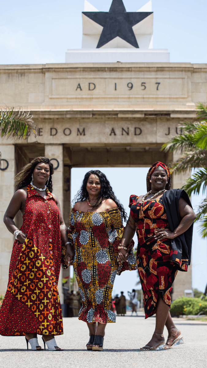 Three women in colorful African dresses standing in front of the Black Star Square monument in Ghana during a heritage tour