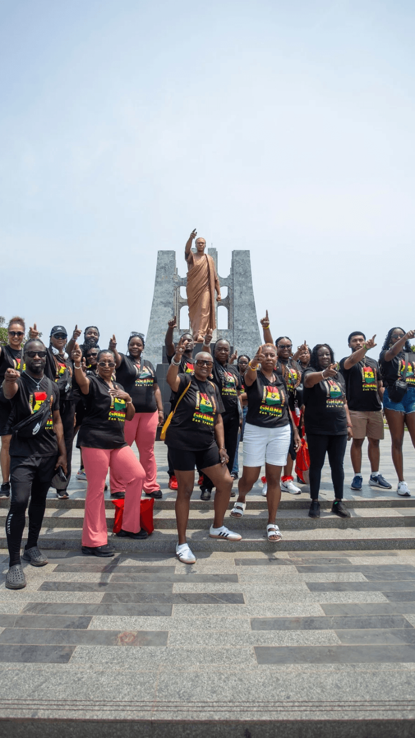A group of touurists standing in front of the Kwame Nkrumah se of a man in orange robes, with many raising their index fingers
