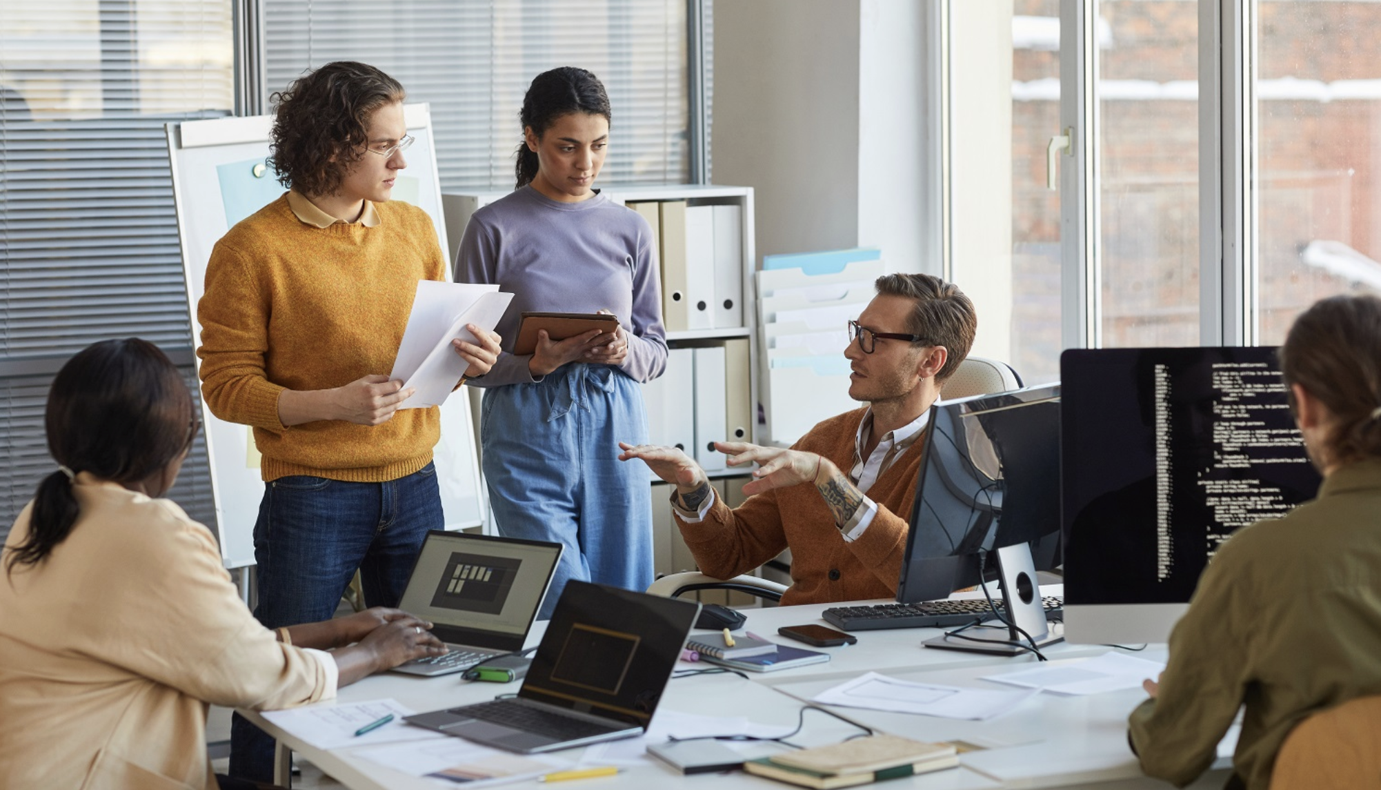 A diverse team of five people working together in a modern office, some standing and some seated at desks with laptops and monitors, discussing a project.