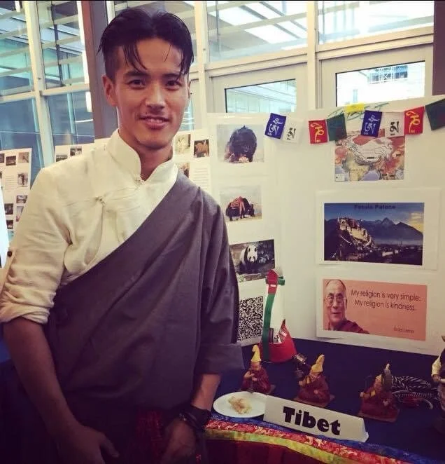 Topten dressed in traditional Tibetan attire standing next to a display booth labeled 'Tibet,' which features images of Tibetan culture, artwork, and a quote from the Dalai Lama about religion and kindness.