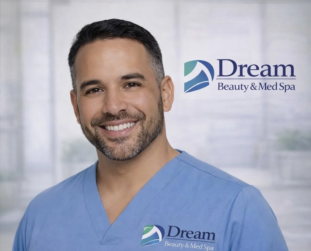A smiling man in blue medical scrubs standing in front of a window with a logo that reads "Dream Beauty & Med Spa".