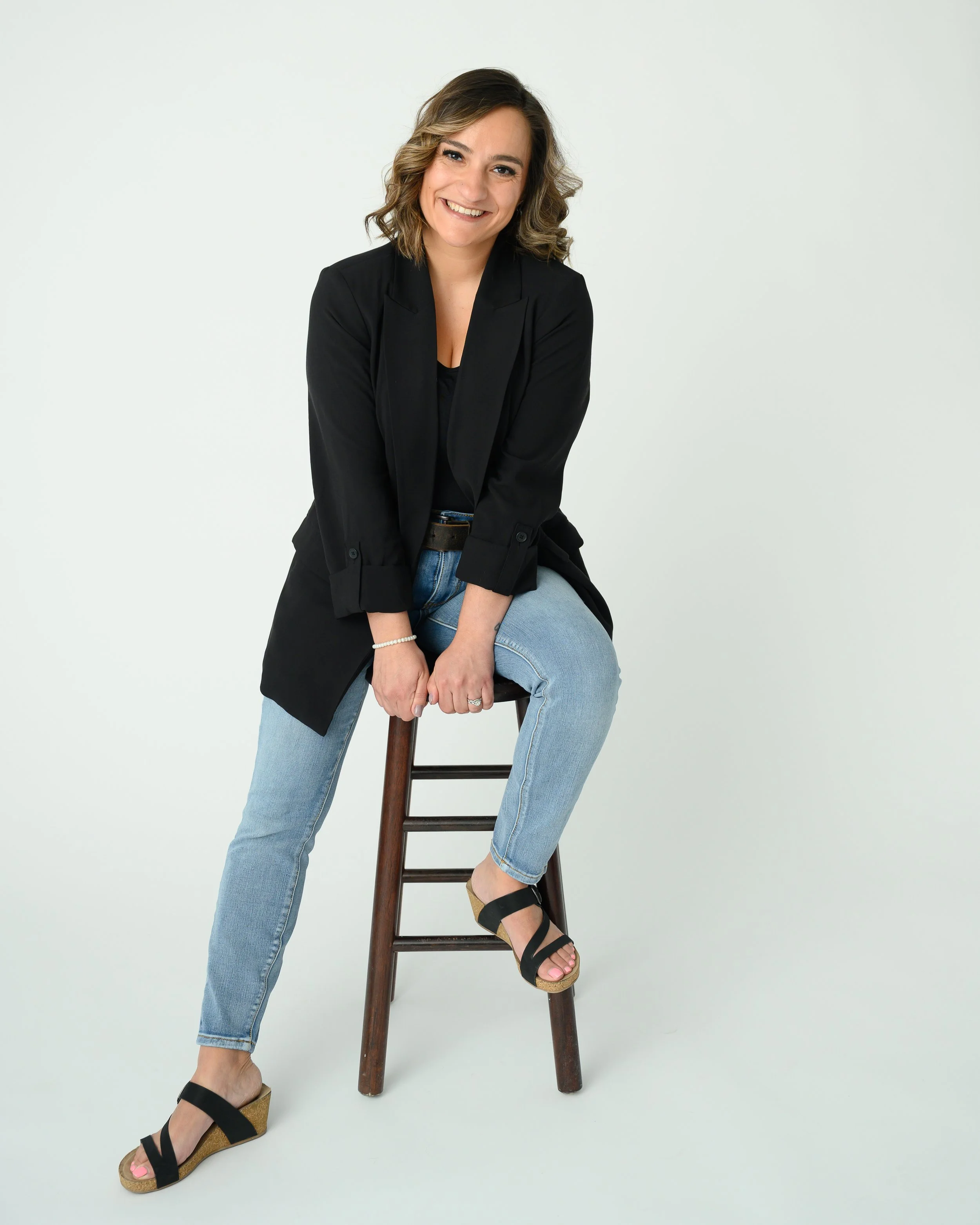 A woman sitting on a wooden stool, smiling, wearing a black blazer, black top, light blue jeans, and black platform sandals, against a plain white background.