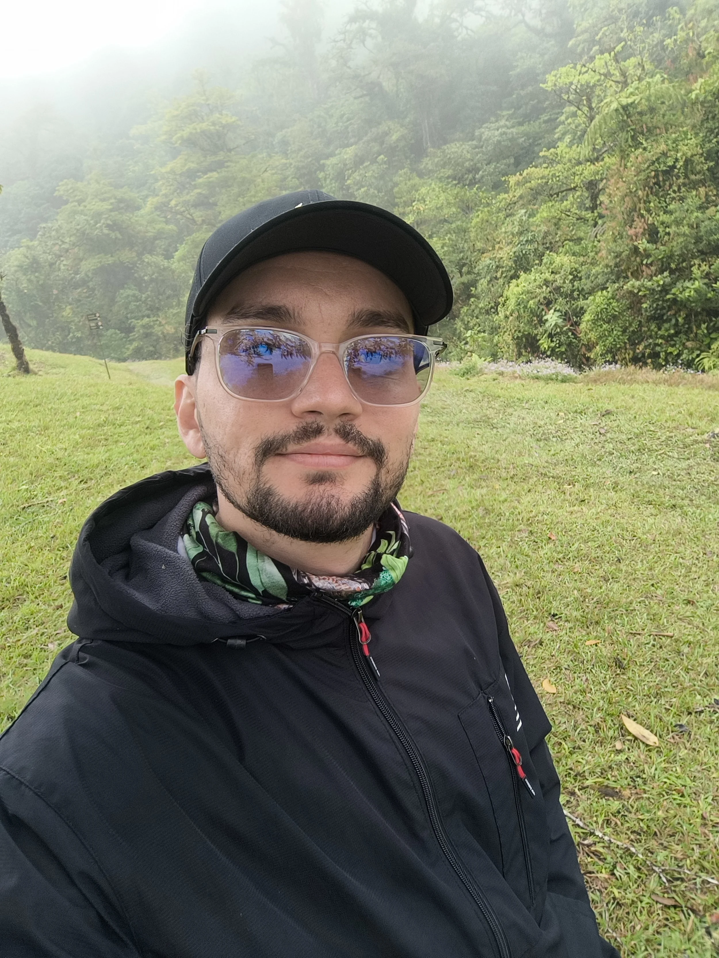 A man with a beard and glasses wearing a black cap and jacket, taking a selfie outdoors in a lush green area with trees and fog in the background.