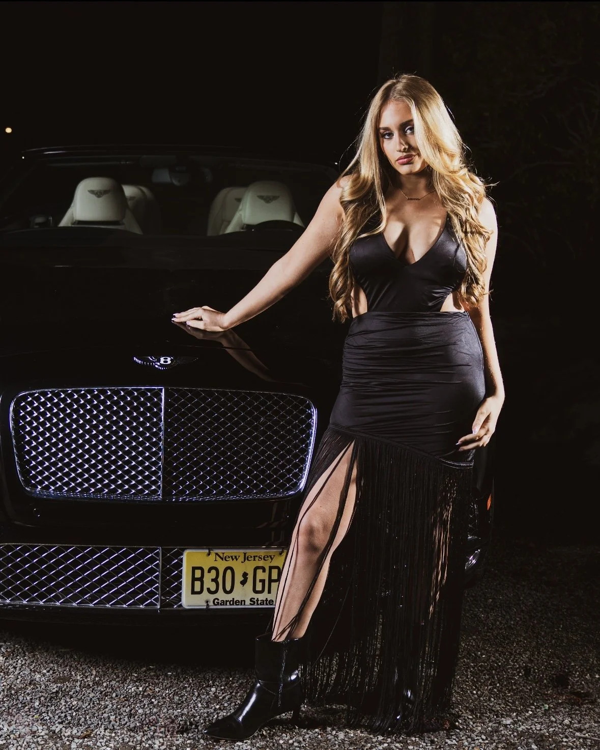 A woman with long wavy hair wearing a black dress with a high slit, standing beside a black luxury car at night.