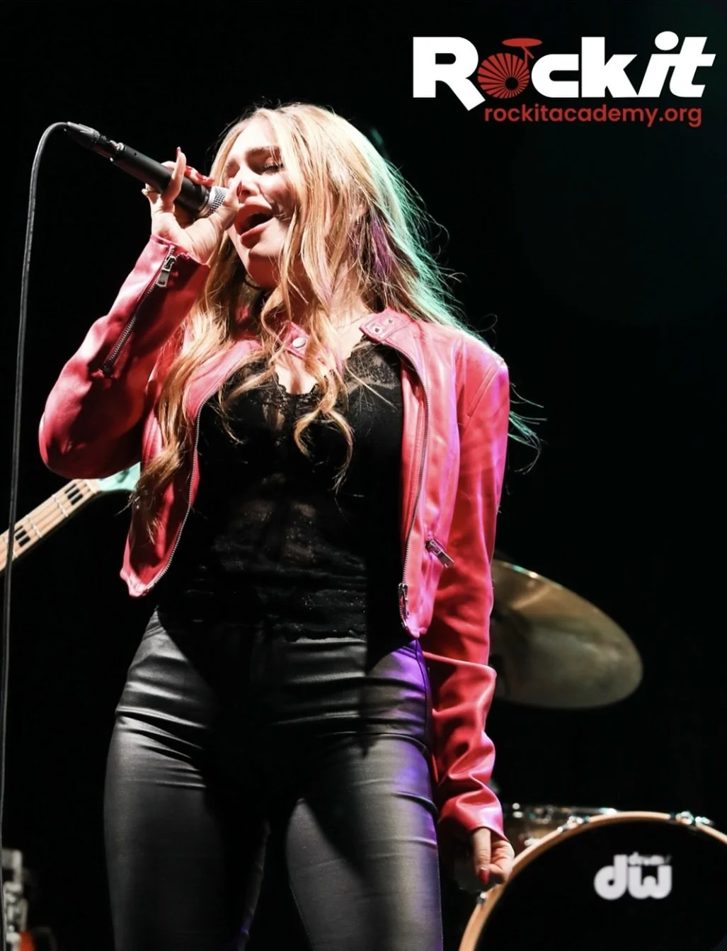 A young woman singing into a microphone on stage, wearing a red leather jacket and black outfit, with a drum in the background. The image has a 'Rockit' logo and website at the top right corner.