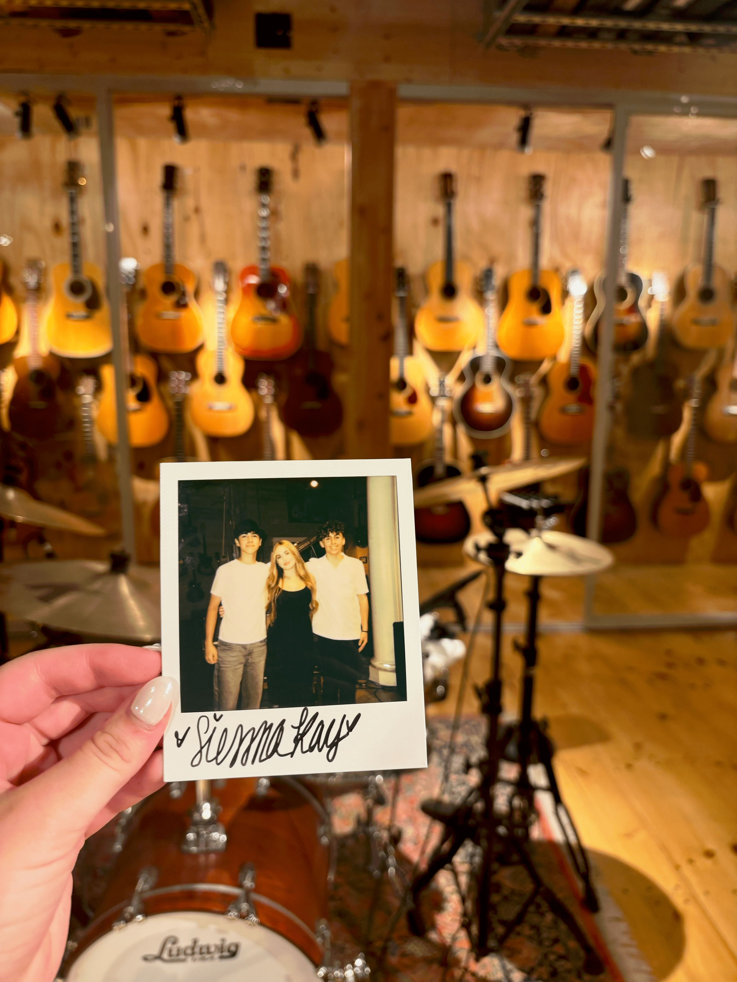 A person holding a Polaroid picture of three individuals in front of a wall of guitars in a music studio. The person holding the photo has light-colored nails.