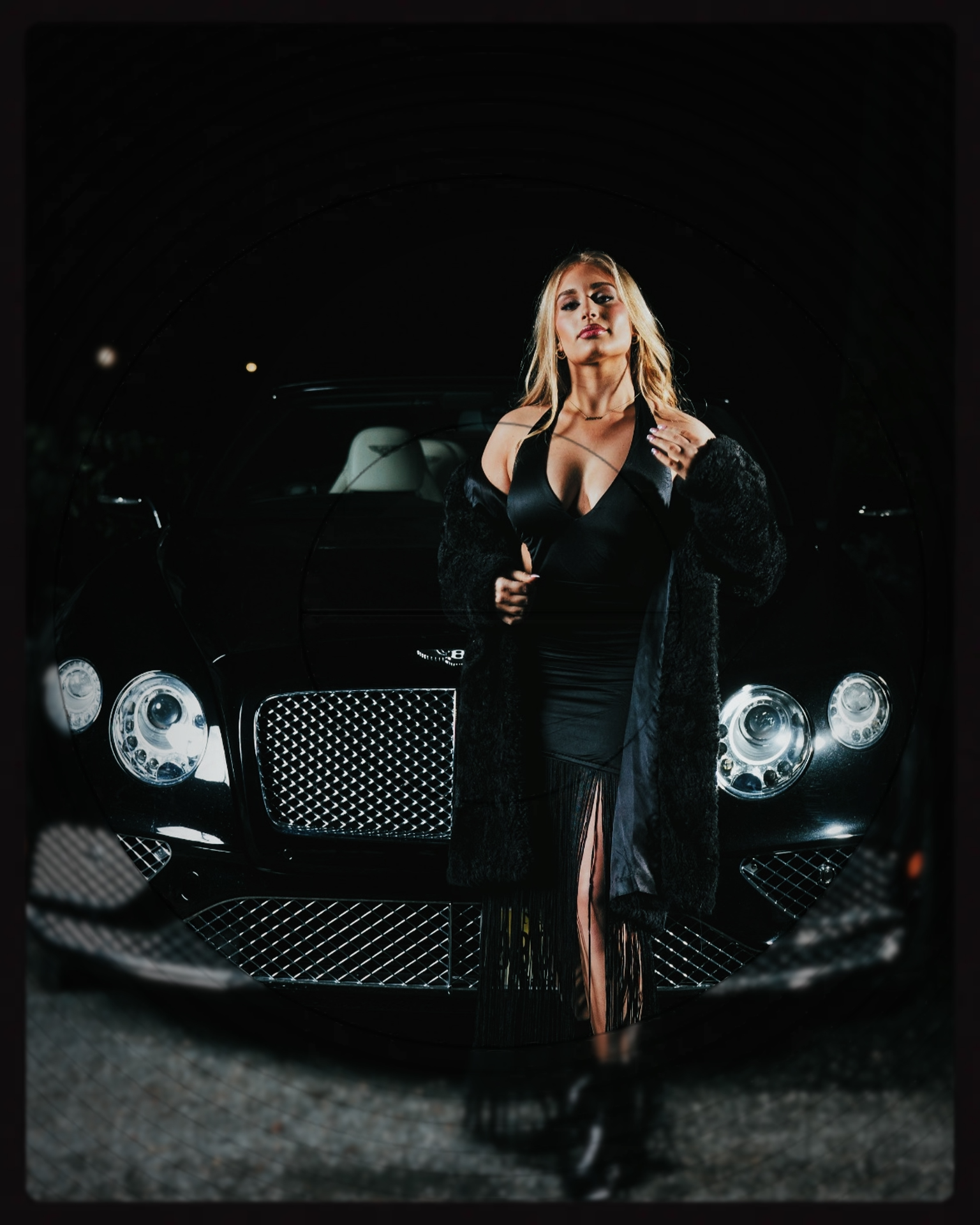 A woman in a black dress and fur coat standing in front of a black luxury car, viewed through a circular mirror.