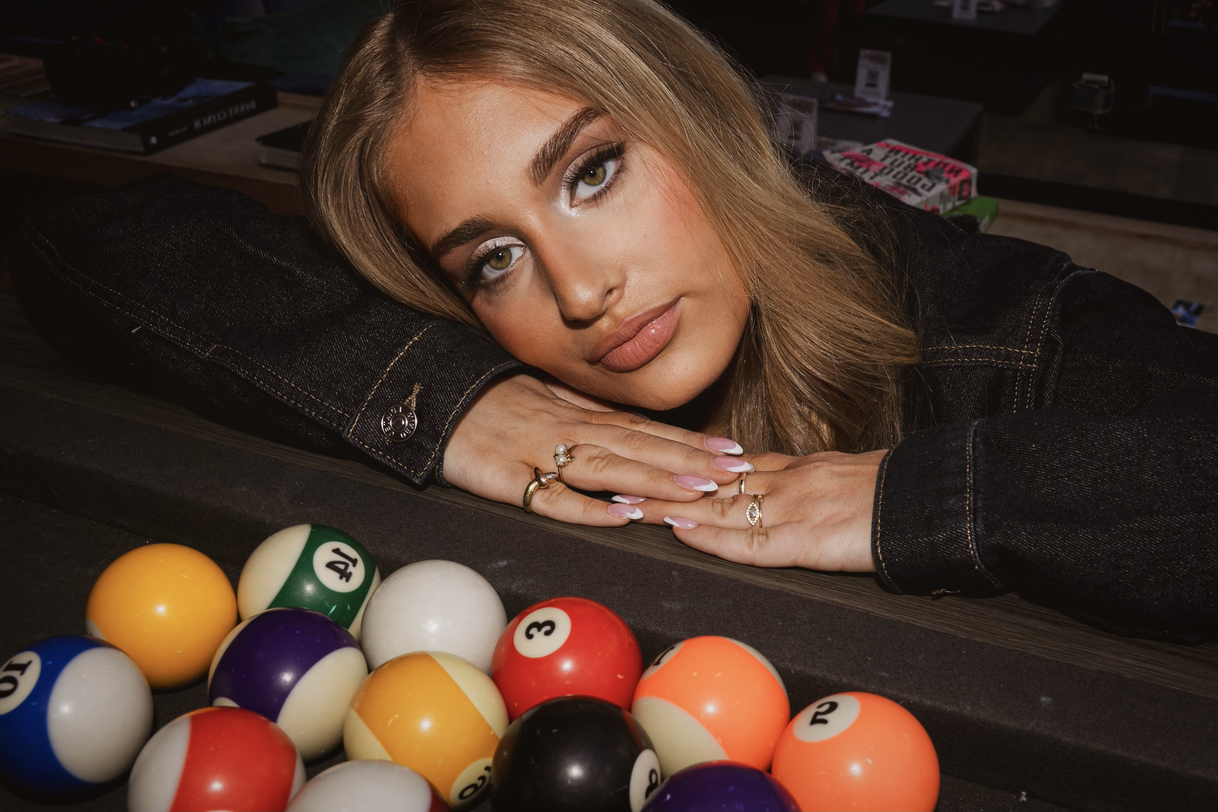 A young woman with blonde hair, wearing a denim jacket and multiple rings, resting her head on her folded arms at a pool table, with colorful billiard balls in the foreground.