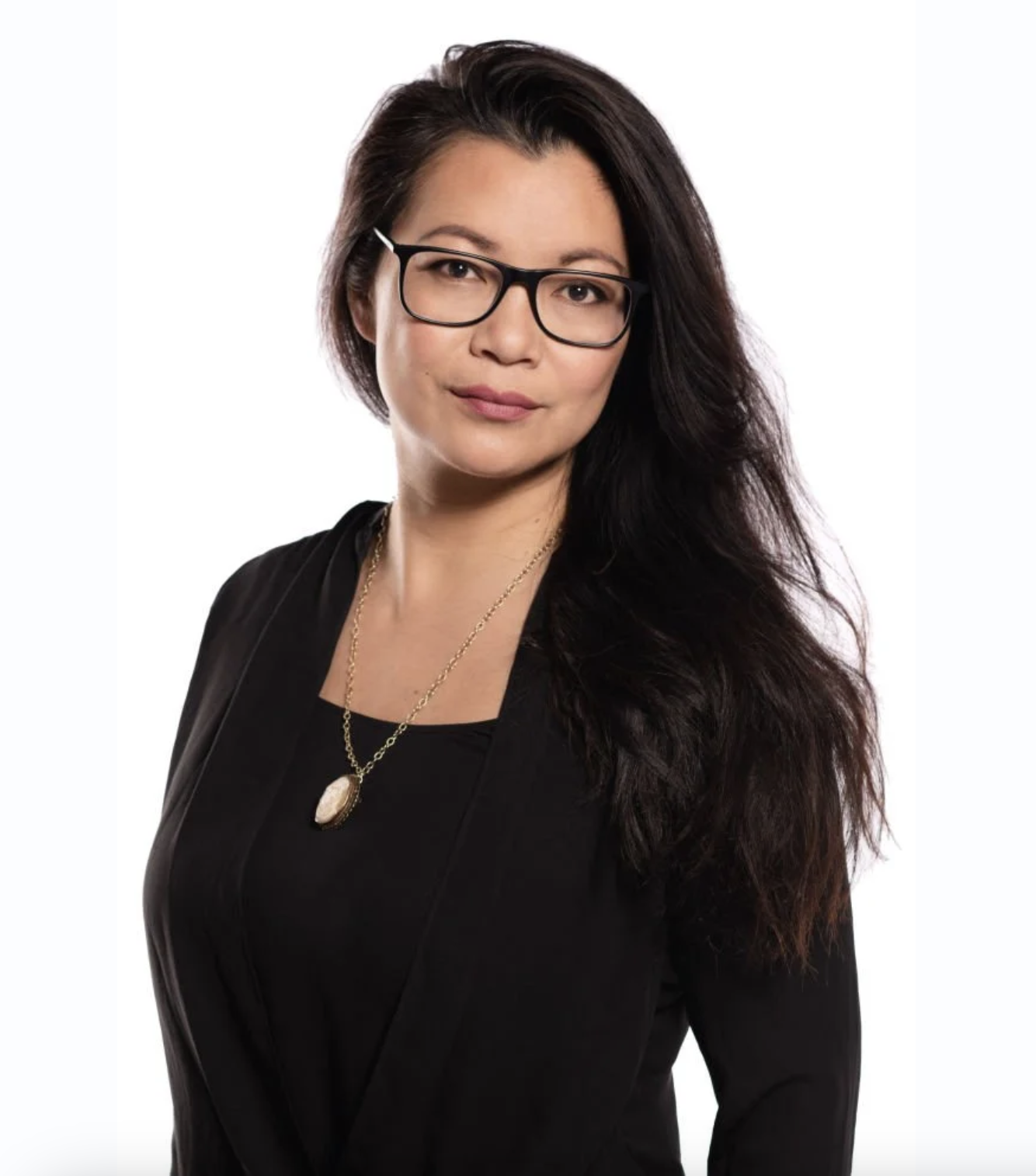 A woman with long dark hair, wearing glasses, a black blazer, and a necklace with a pendant, posing against a white background.