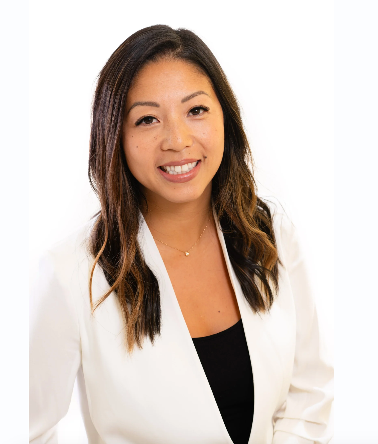 A professional woman with long dark brown hair styled in loose waves, wearing a white blazer over a black top, smiling and looking at the camera against a plain white background.