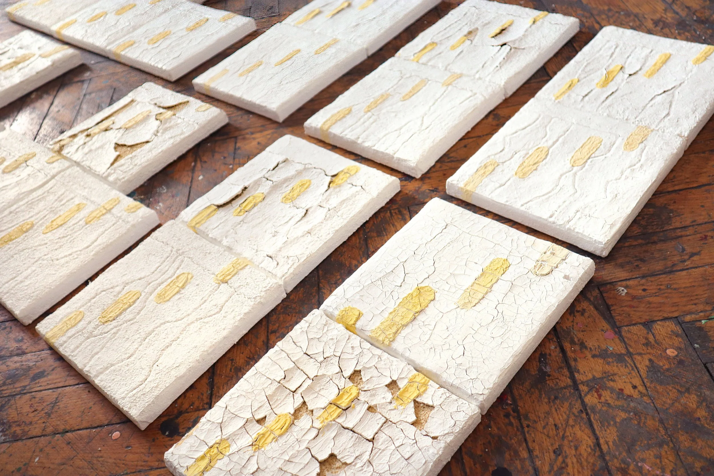Multiple white textured tiles with cracks and yellow designs laid on a wooden surface.