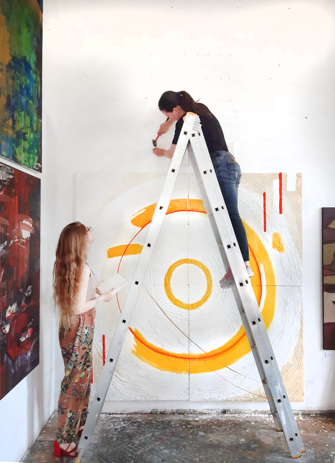 Two women working on an art show setup, with one standing on a ladder and the other on the ground holding a small artwork to hang on the wall in an assembly. Below is an artwork featuring 2 linear yellow circles, on a white textured background.