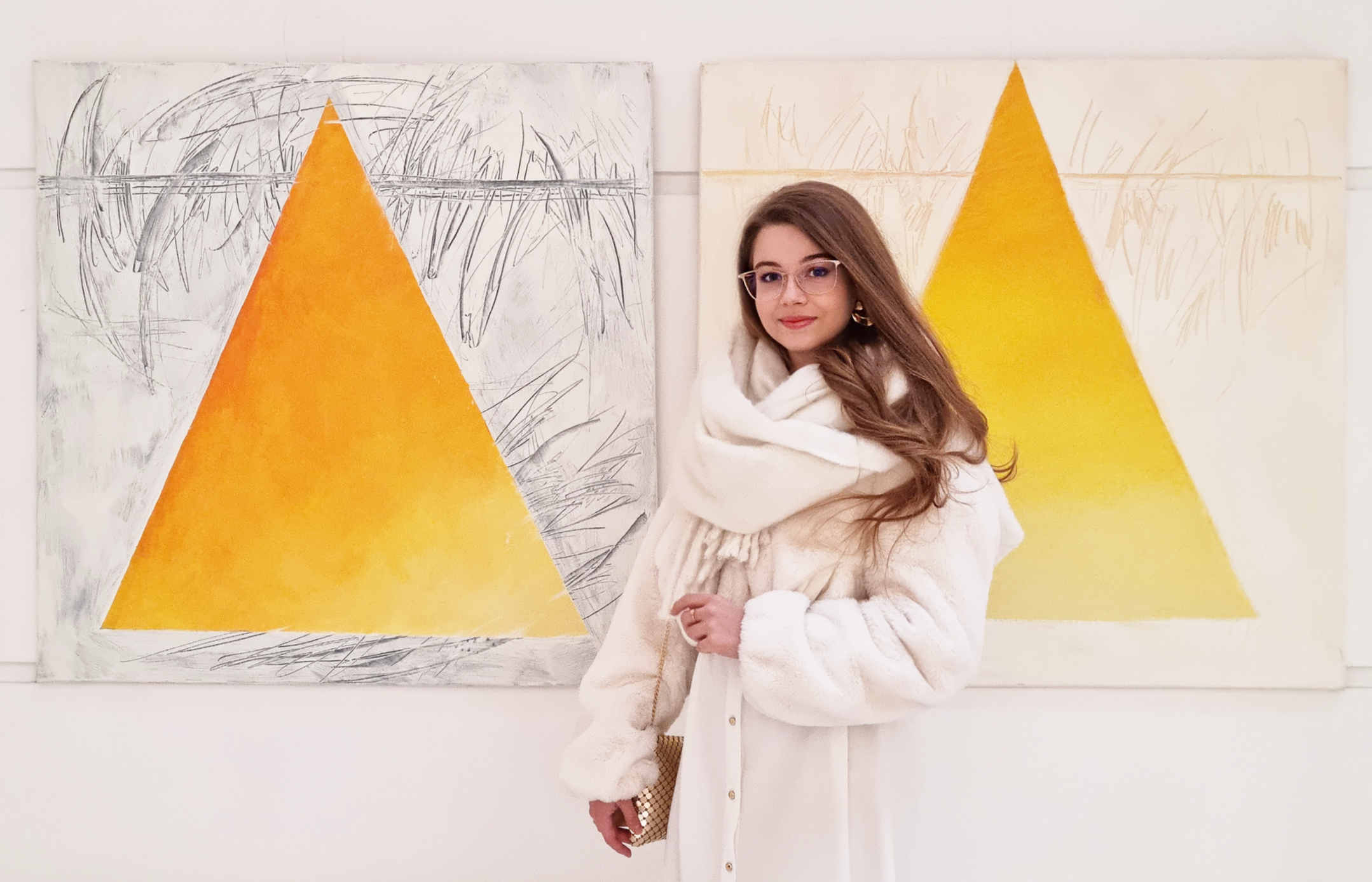 The artist, a young woman with long, dark-blonde hair and glasses standing in front of her two large abstract paintings with yellow geometric triangles.