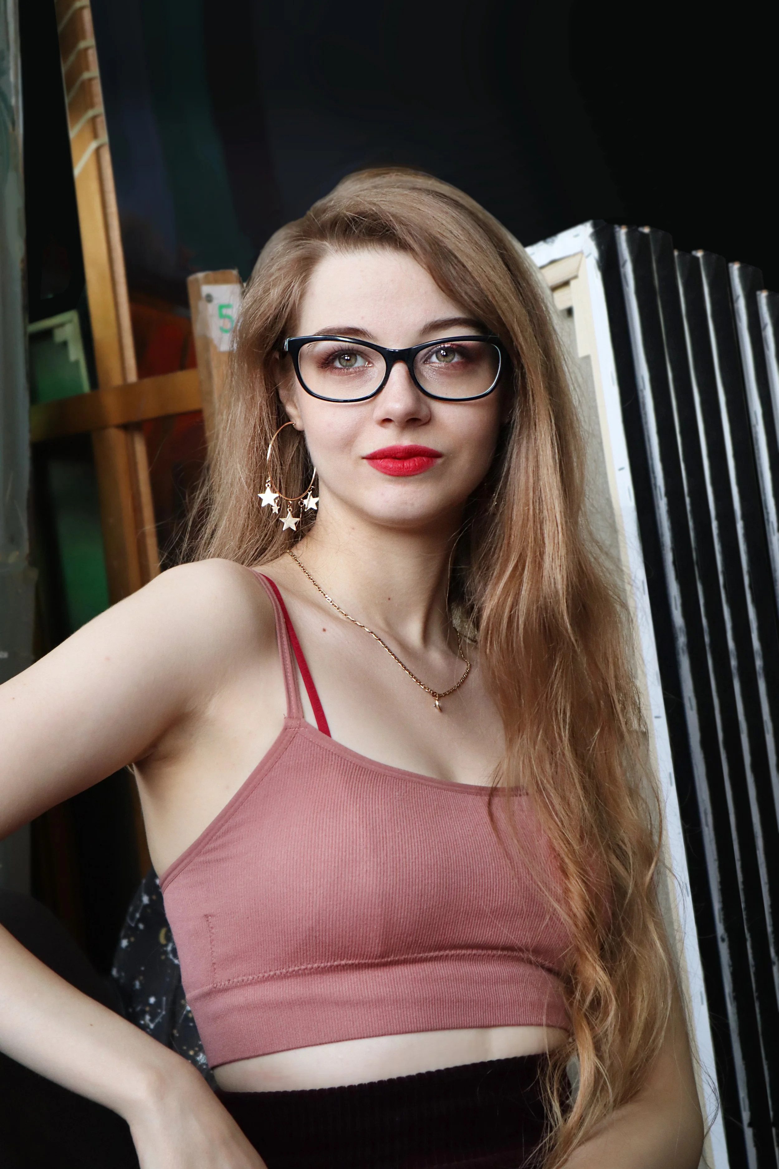 Andeea-Eliza Petrov, a young woman with long, wavy, dark-blonde hair wearing glasses, star-shaped earrings, a gold chain necklace, a pink tank top, and black skirt, in front of a background with an easel and canvases.