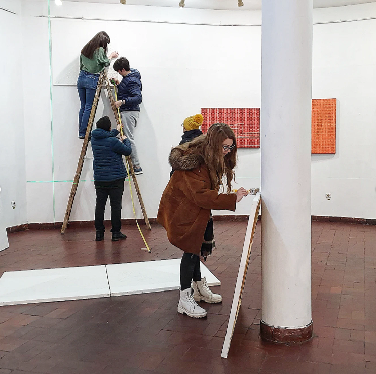 People in an art gallery working on an interactive art piece, with some on a ladder and others painting on a wall or standing nearby.