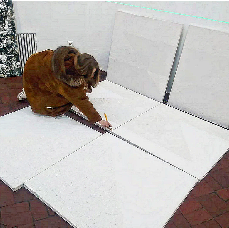 Person in brown coat kneeling and marking white tiles with a yellow marker on a floor.