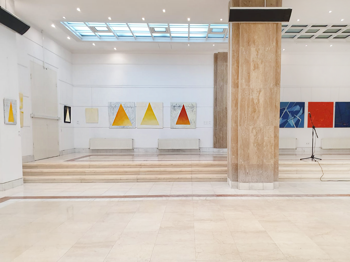 Art gallery with paintings on white walls, a small stage with steps, a microphone, and a large skylight ceiling.