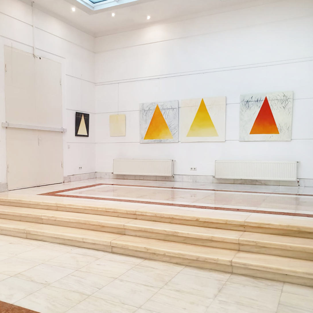 An art gallery with white walls and marble flooring, displaying five abstract paintings featuring triangular shapes in shades of orange, yellow, and red.