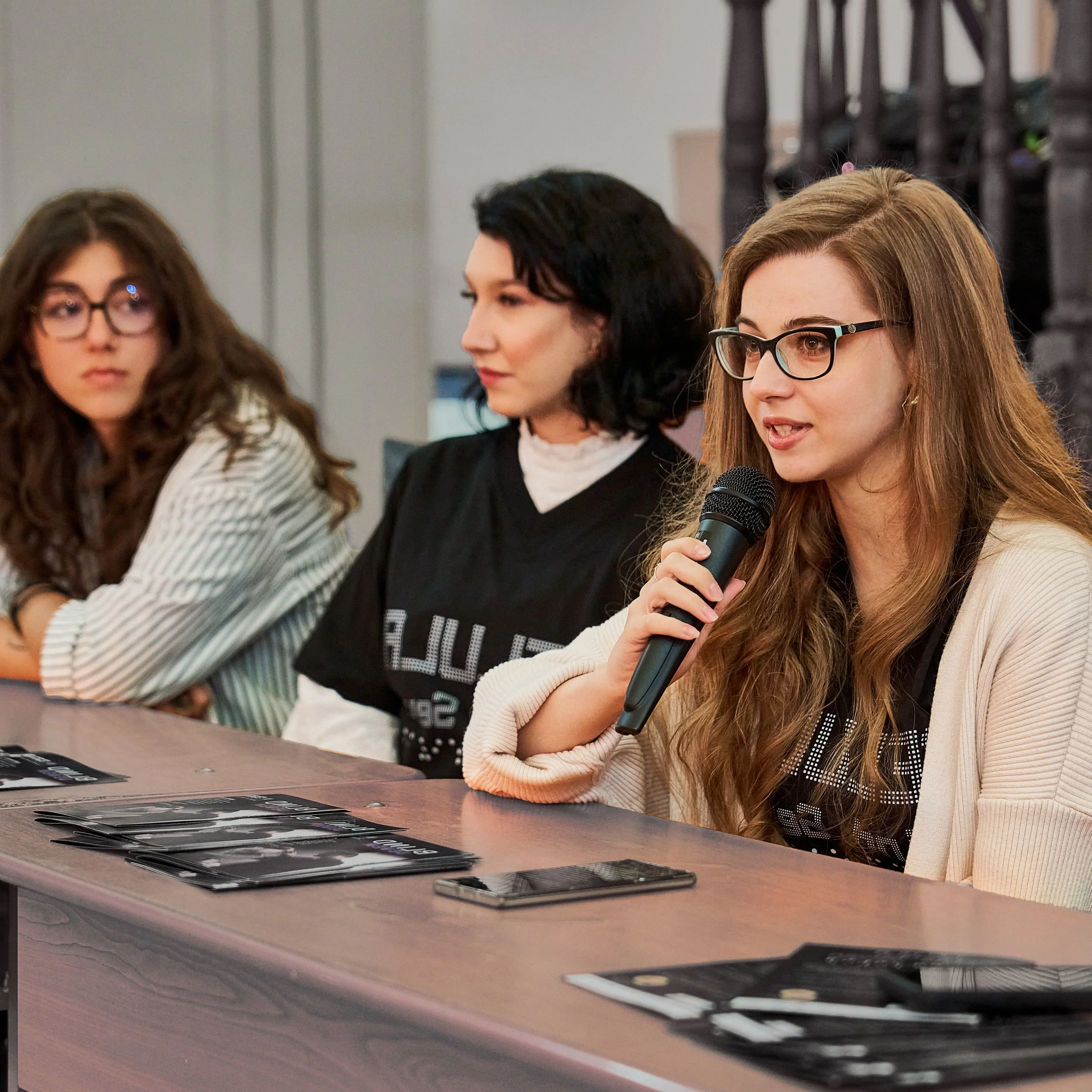 Three curator moderating a q&a session, speaking into a microphone alongside two of the artists involved in the Blind Seven art project, seated at a table with flyers in front of them.