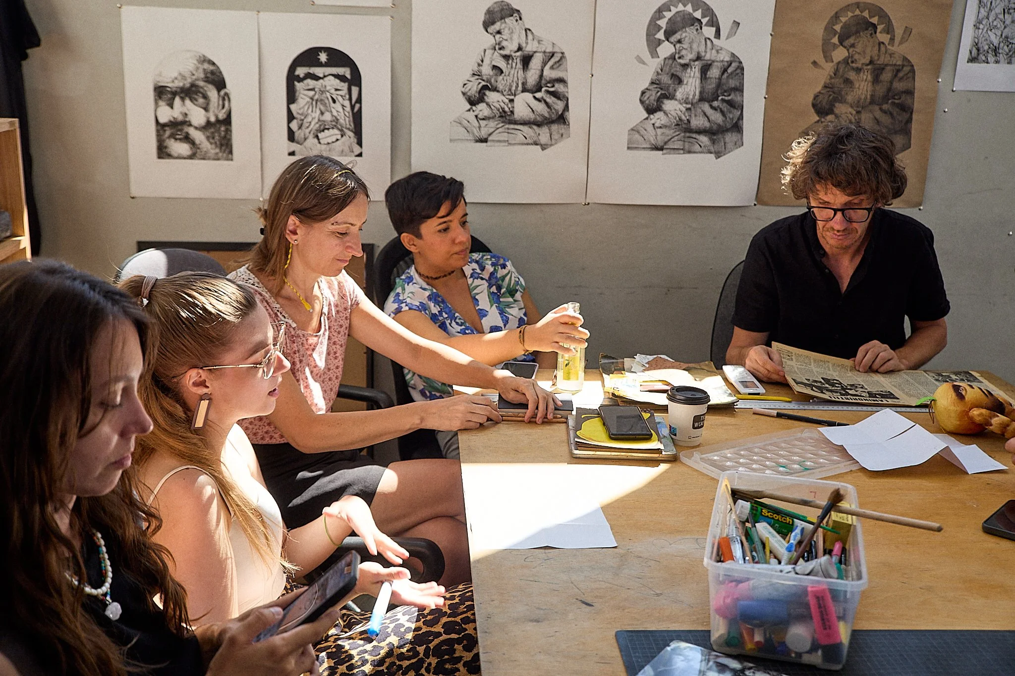 A group of five women artists and one man, sitting at a table at the graphic arts university studio in Wroclaw, Poland, with artworks displayed on the wall behind them. They are engaged in an exchange of ideas, surrounded by art supplies and papers.