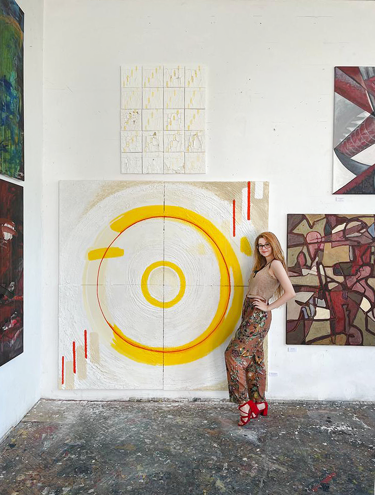 A woman with blonde hair, glasses, a beige sleeveless top, wide colorful patterned pants, and red high heels stands at her university semester exhibition, posing next to her large abstract painting on the wall featuring huge bullseye yellow circles.