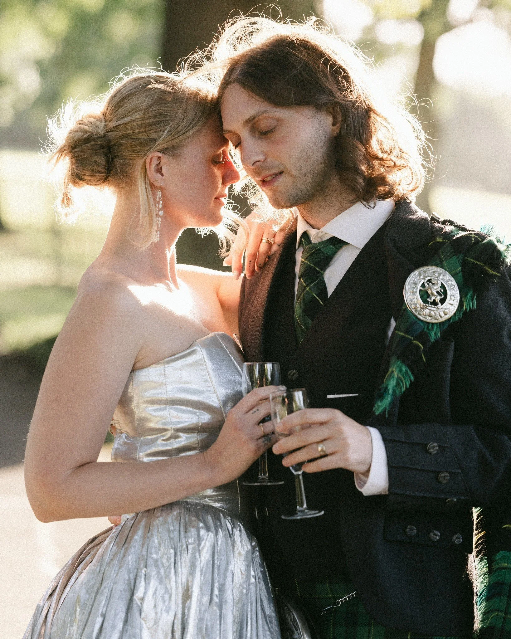 A couple is shown in a close, intimate pose outdoors at sunset, with their foreheads touching and eyes closed. The woman wears a strapless, metallic silver dress and elegant jewelry, while the man is dressed in a traditional Scottish outfit, includin