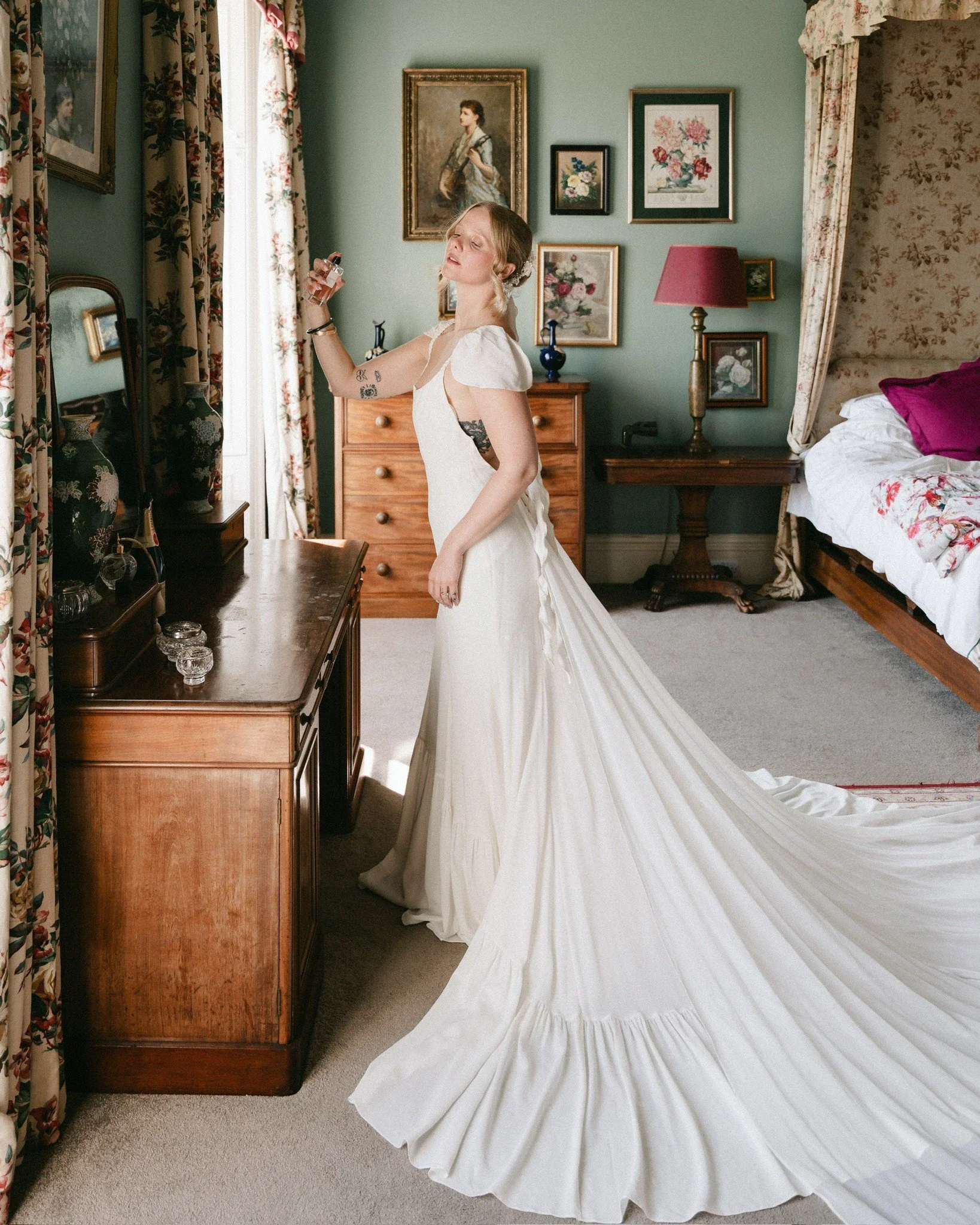 A woman in a white wedding dress holding a perfume bottle in a vintage bedroom with floral and portrait artwork, wooden furniture, and floral curtains.