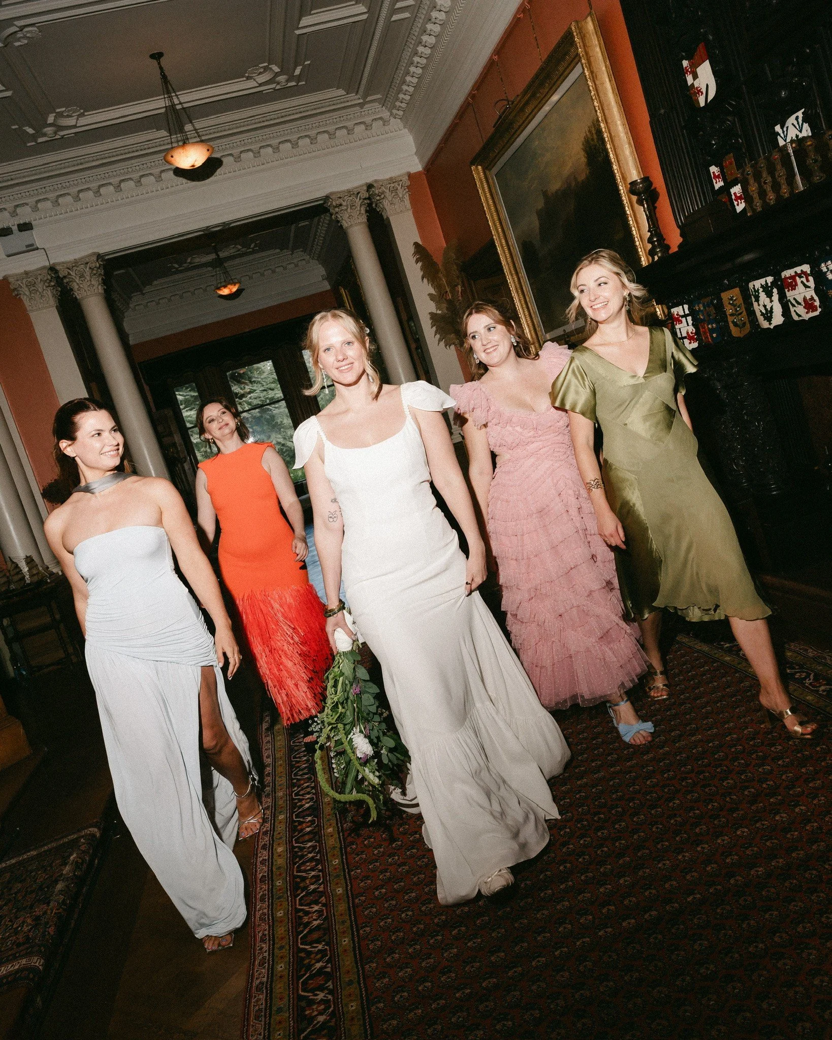 A group of women dressed in elegant formal dresses walking indoors in an ornate room with classical architecture and paintings.