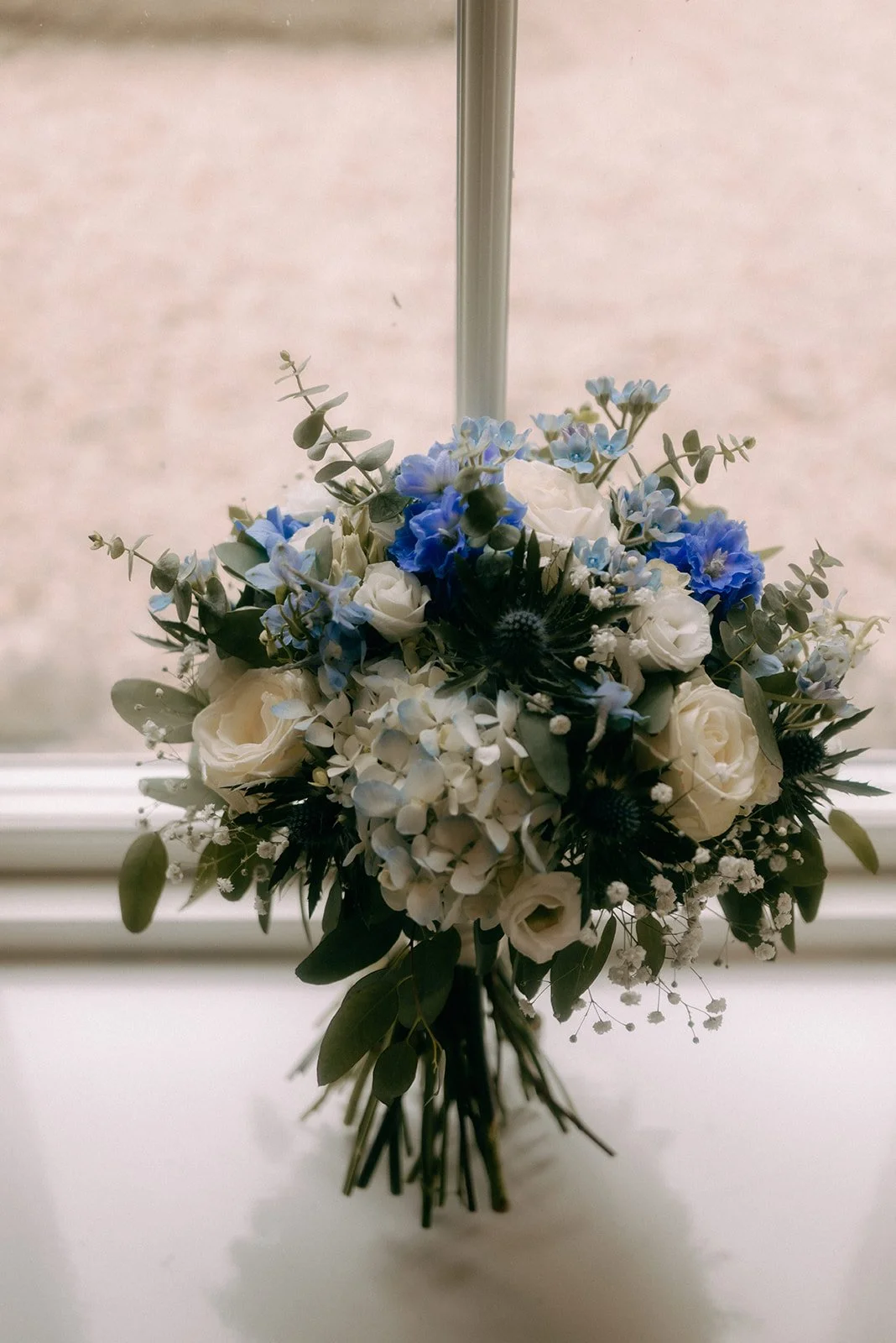 A bouquet of white roses, blue hydrangeas, and greenery, hanging upside down in front of a window.