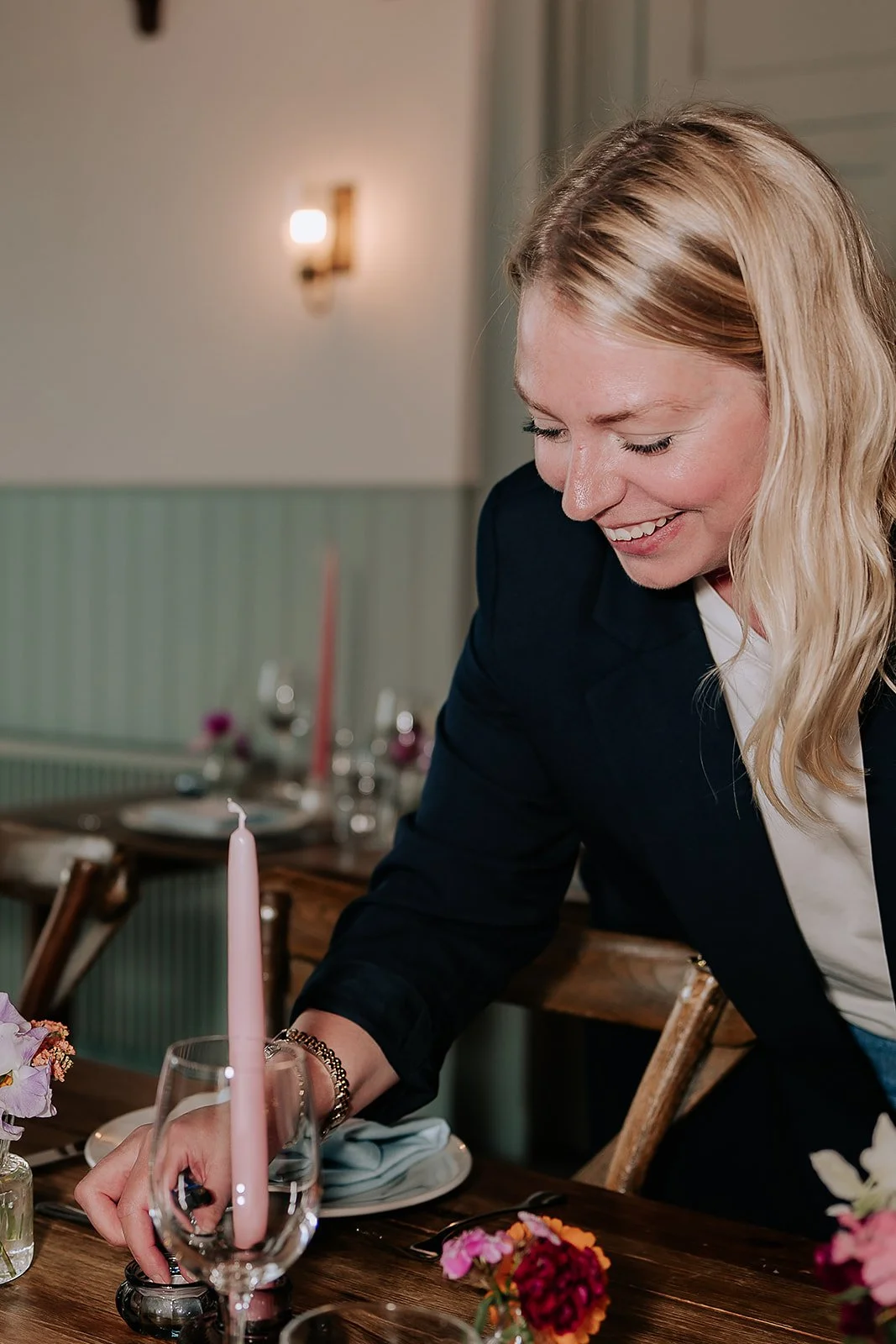Woman smiling and decorating a dining table with flowers and a pink candle in a glass holder.
