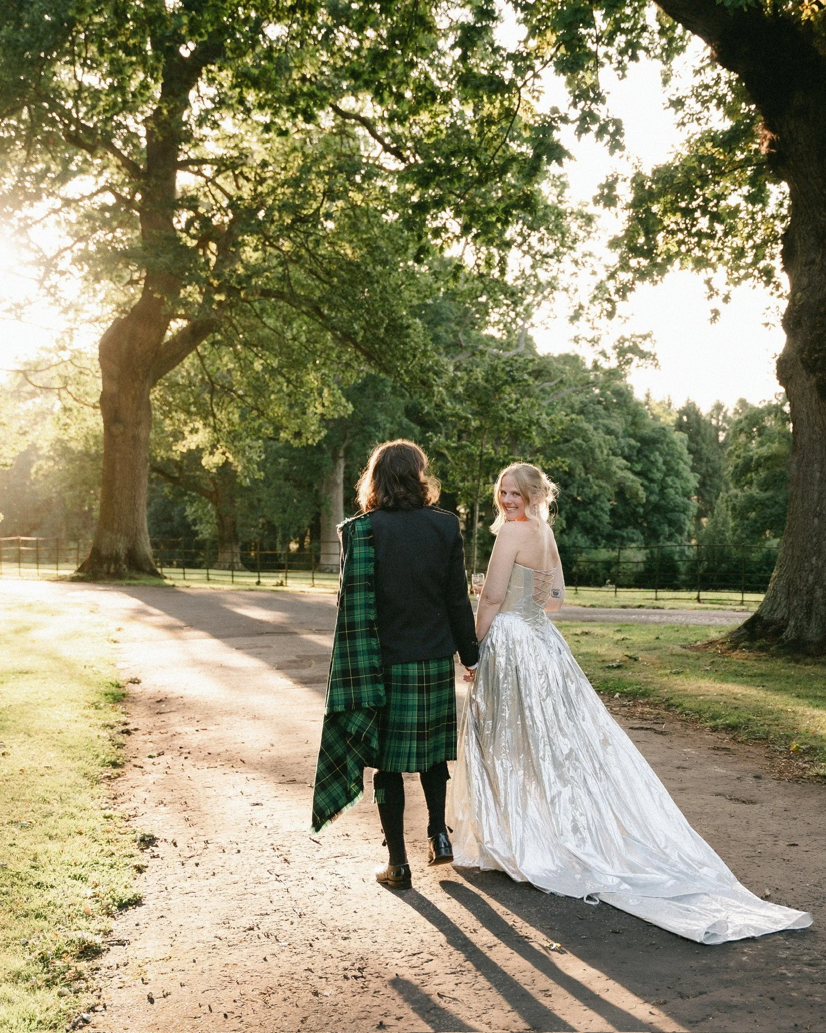 Two women walking away from the camera on a dirt path in a park, one in a white wedding dress and the other in a green and black tartan coat, holding hands. Tall trees with green leaves surround them, with sunlight filtering through.