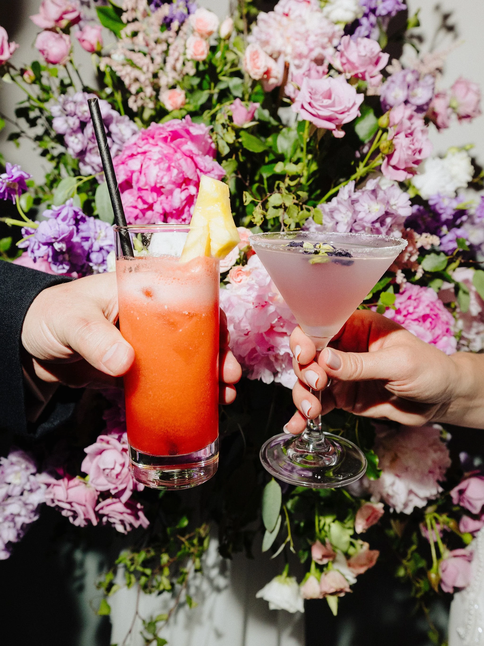 Two hands holding colorful cocktails in front of a vibrant floral background.