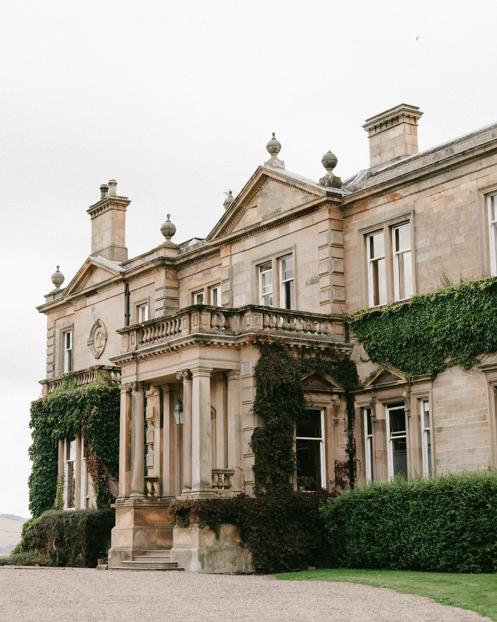 A historic stone mansion with classical architectural features and ivy growing on its walls, surrounded by a gravel pathway and greenery.