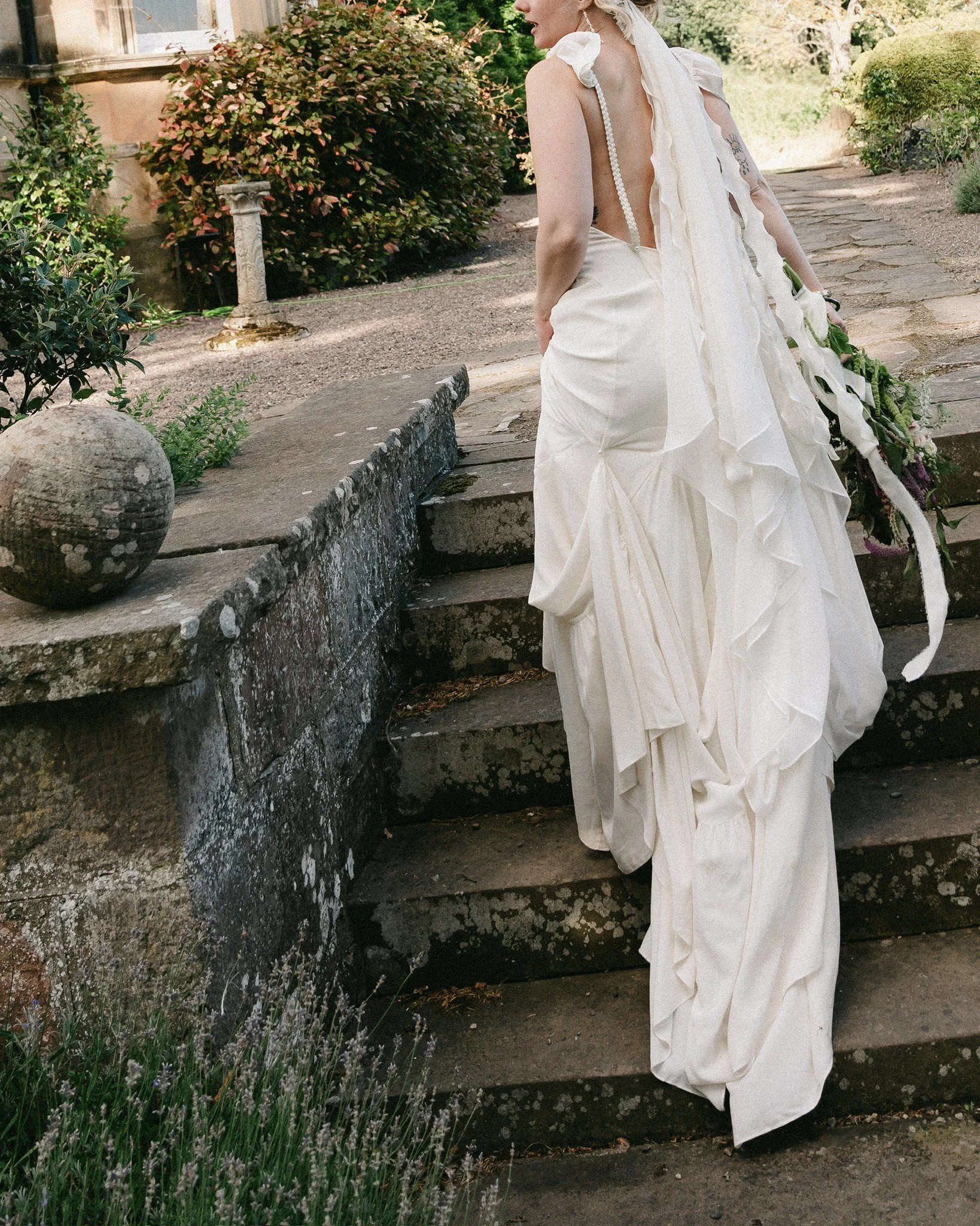 A woman in a white wedding dress standing outdoors on stone steps, holding a bouquet, with greenery and flowering plants around.