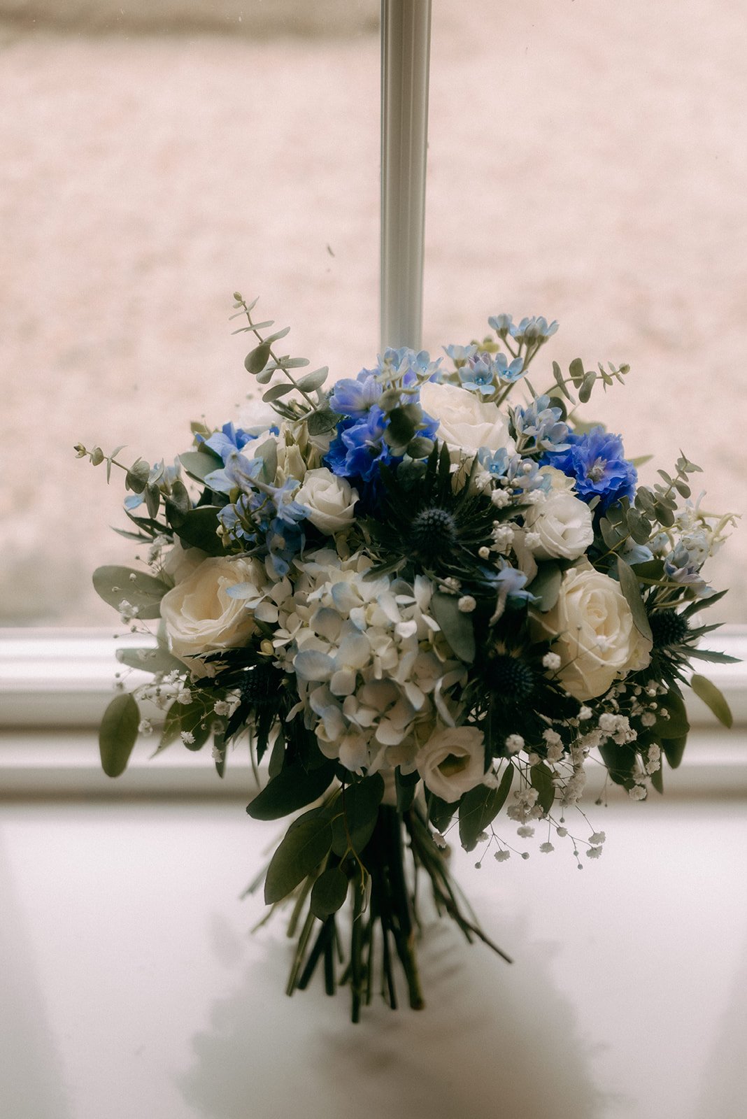 A bouquet of white, blue, and green flowers hanging upside down in front of a window.