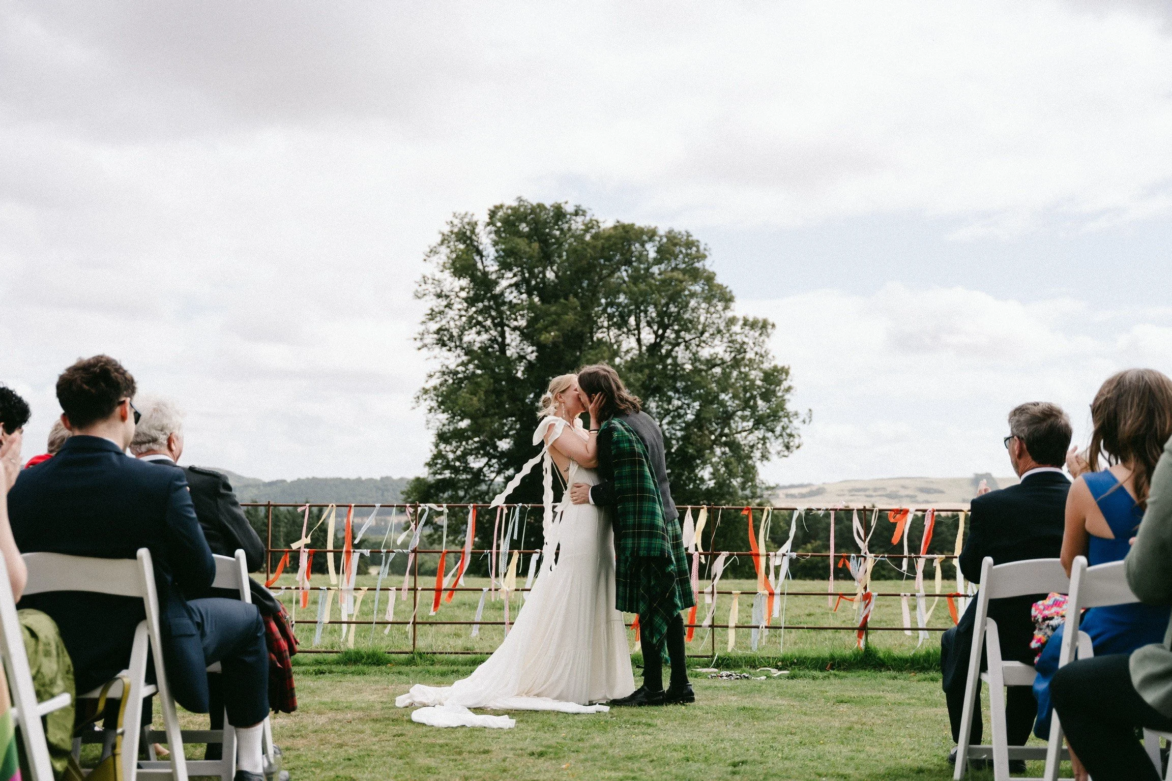 A couple is kissing at an outdoor wedding ceremony. The bride is wearing a white dress with a train, and the groom is dressed in a green plaid kilt. Guests are seated on white chairs, watching the couple, with a scenic countryside and a large tree in