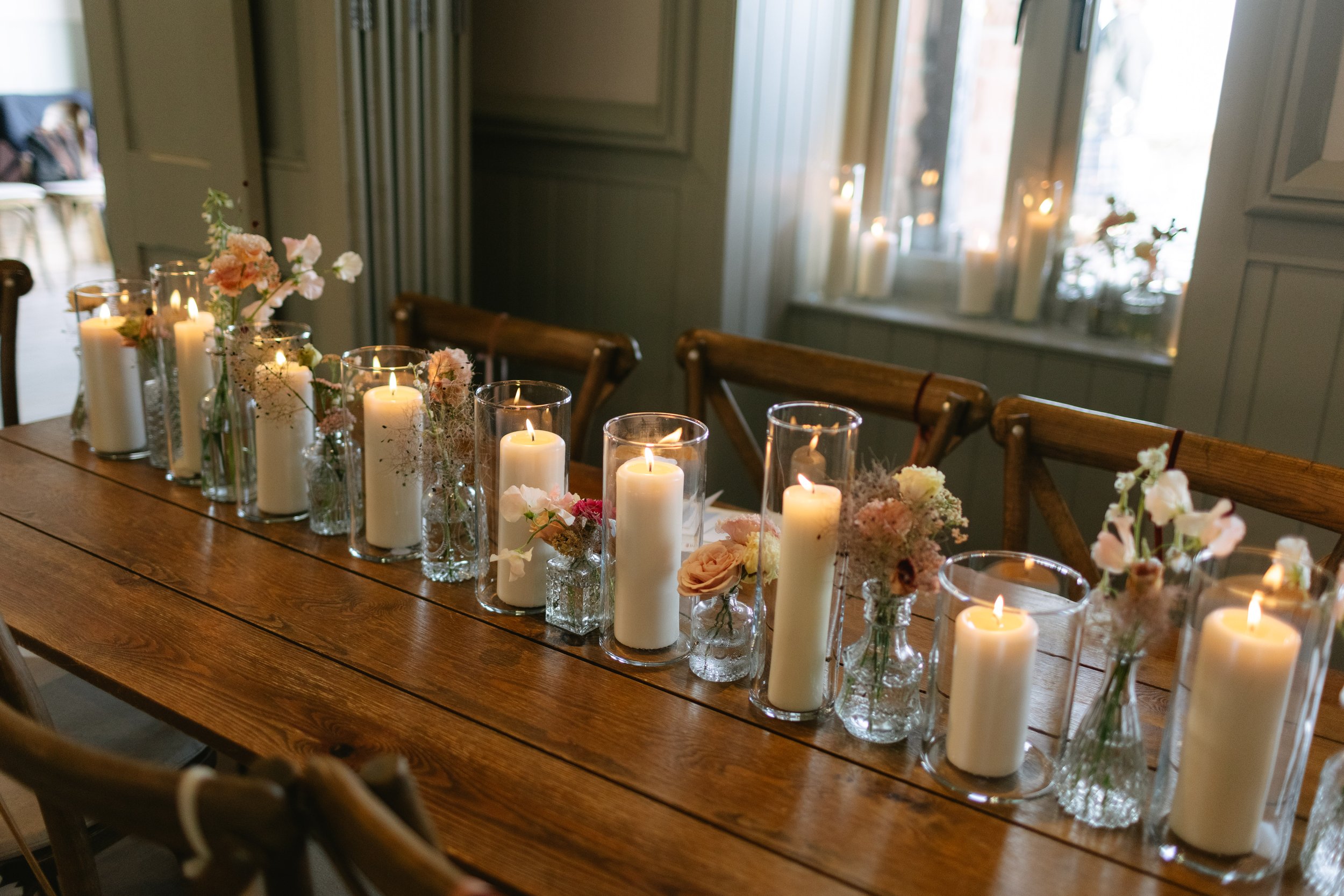 A long wooden dining table decorated with white candles in glass holders and pink and white floral arrangements in clear vases, set in a cozy, softly lit room with large windows and curtains.