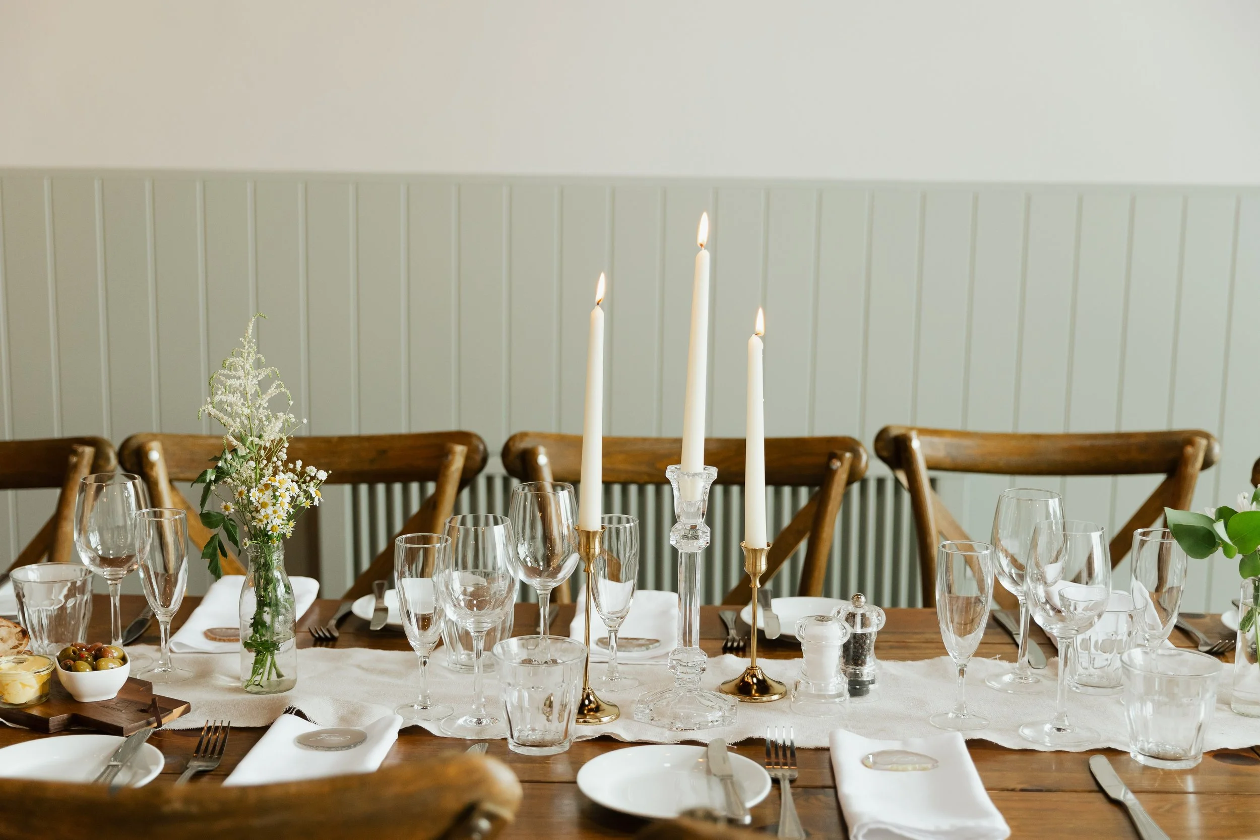 A dining table set for a special occasion with elegant glassware, white candles, and floral arrangements. Wooden chairs are positioned around the table against a light-colored wall with vertical paneling.