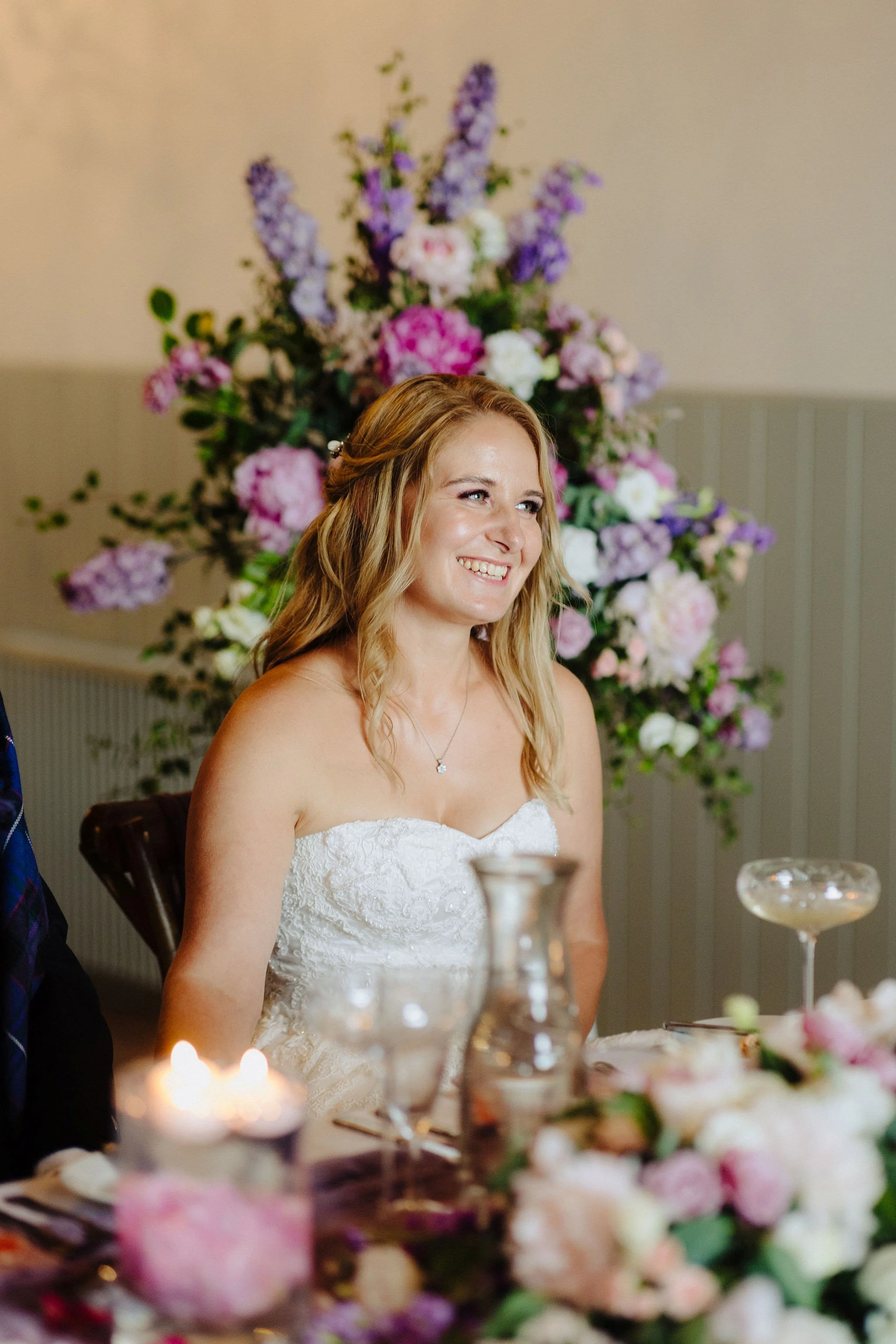 A woman smiling at a wedding reception, seated at a table decorated with flowers, candles, and wine glasses, with a large floral arrangement in the background.