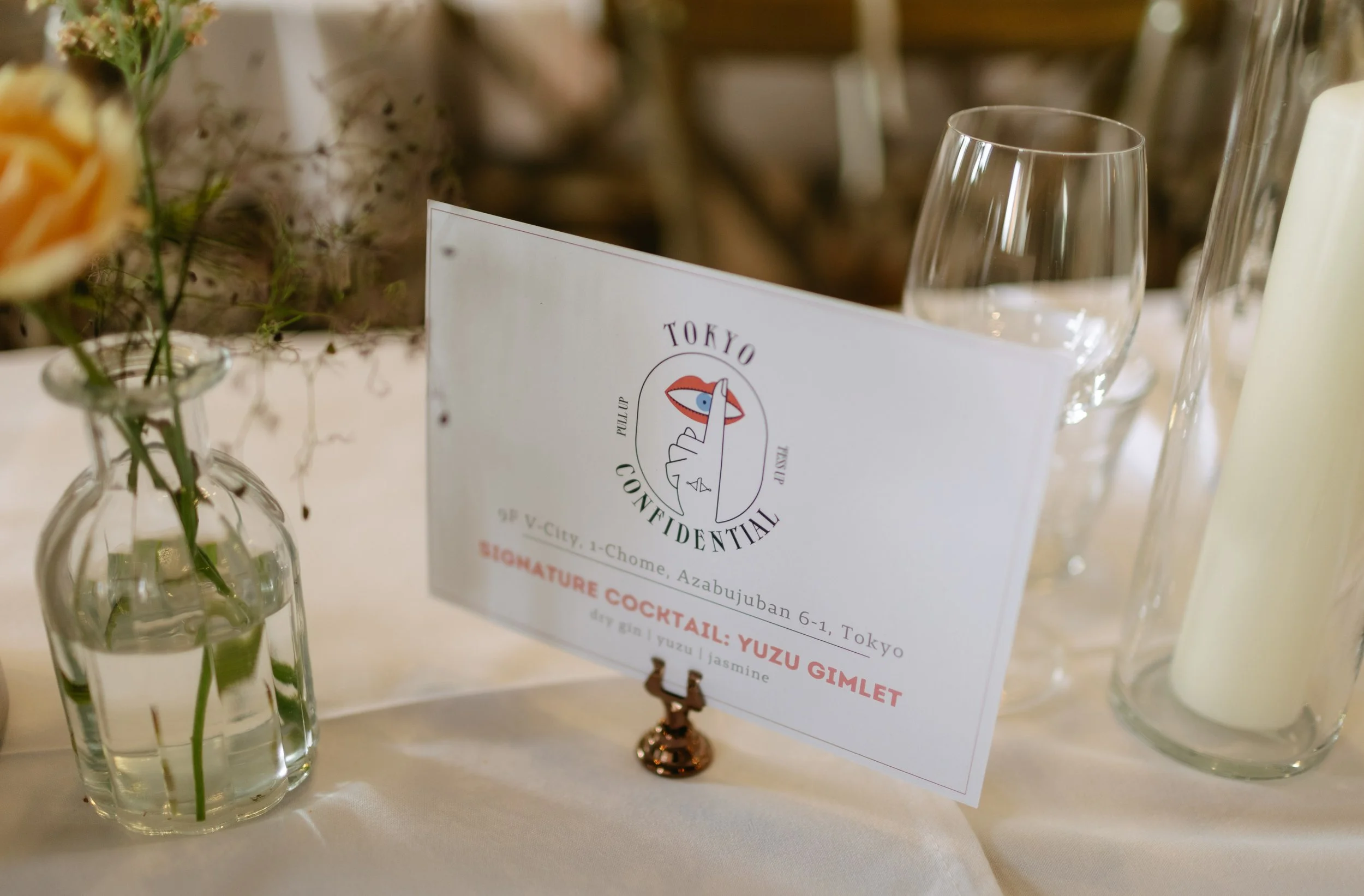 A table setting with a small bouquet of flowers in a glass vase, a pair of empty wine glasses, and a menu card on a white tablecloth.