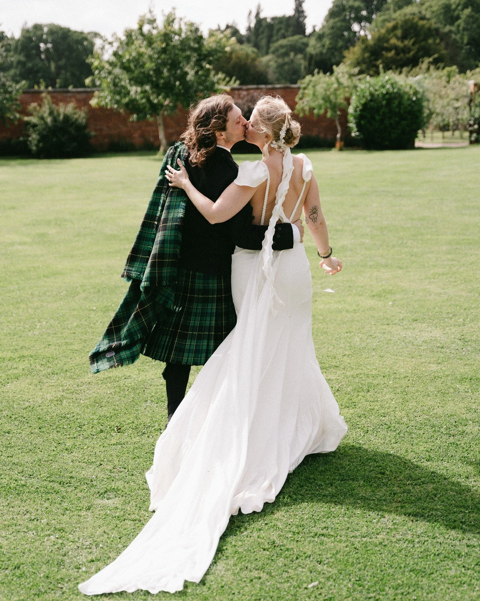 A couple kissing outdoors on a grassy field, the woman wearing a white wedding dress and the man wearing traditional Scottish attire with a tartan kilt and jacket.