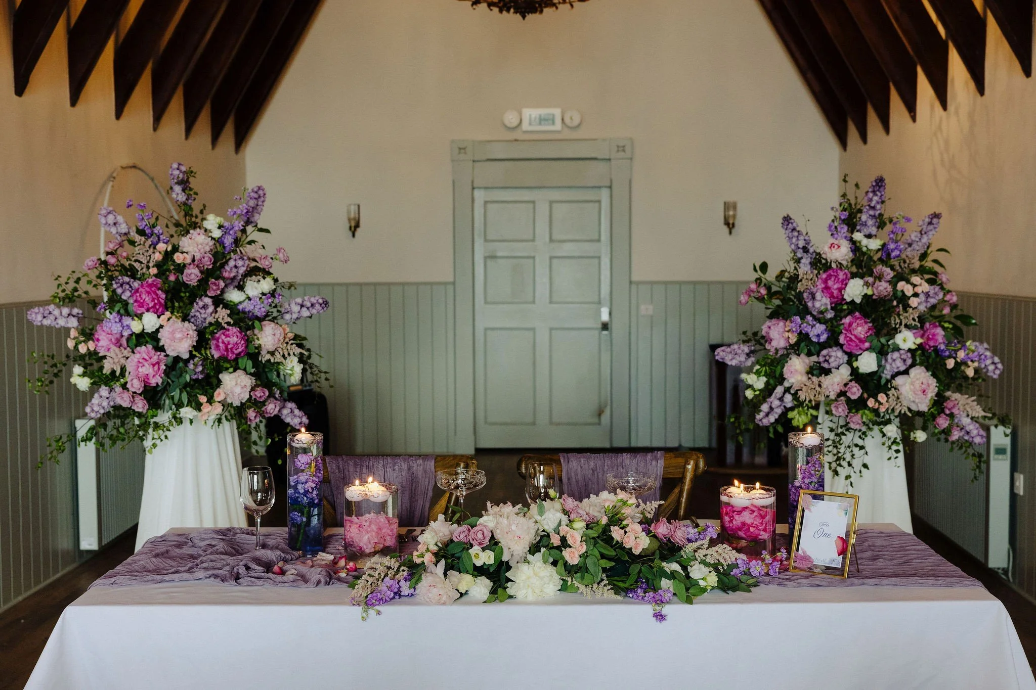 A wedding reception table decorated with large floral arrangements of pink, purple, and white flowers, candles, and a framed sign reading 'Table One' on a white tablecloth, with a background of beige walls and dark wooden ceiling beams.