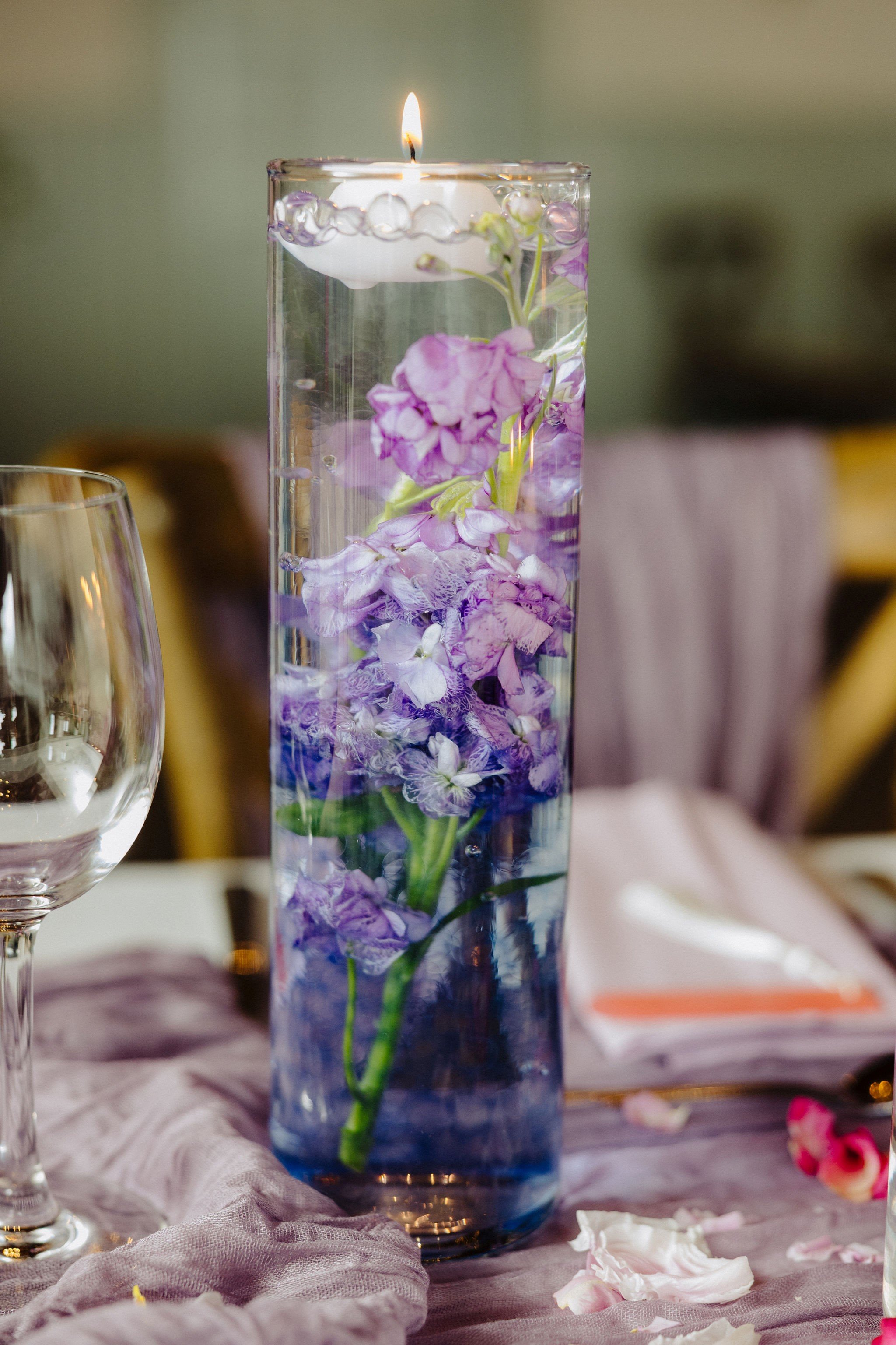 A tall glass vase with purple flowers submerged in water, with a floating candle on top, on a table with scattered rose petals and a pink cloth.