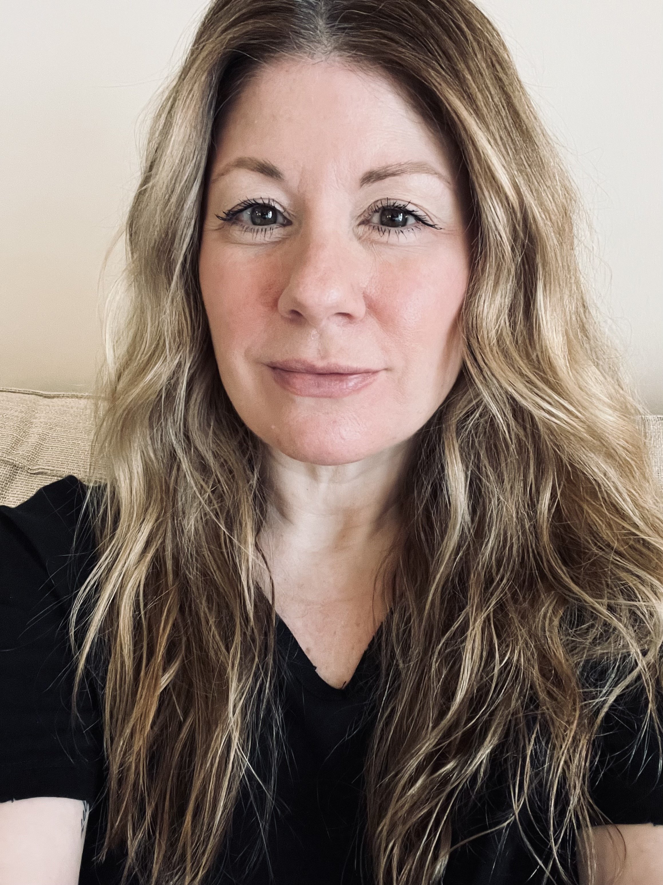 A woman with long, wavy blonde hair and light hazel brown eyes, wearing a black top, sitting on a beige couch in front of a plain white wall.