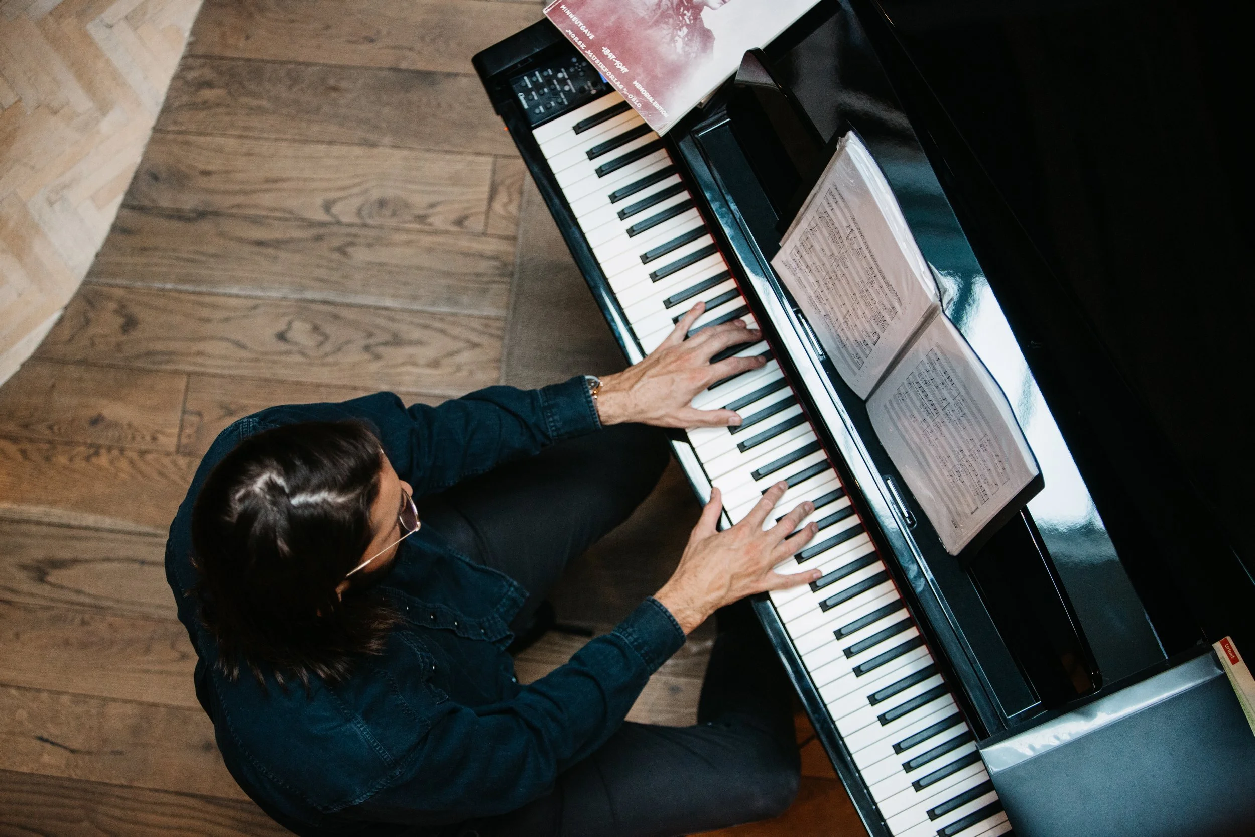 A person playing a black grand piano with sheet music and an electronic device on top, seen from above.