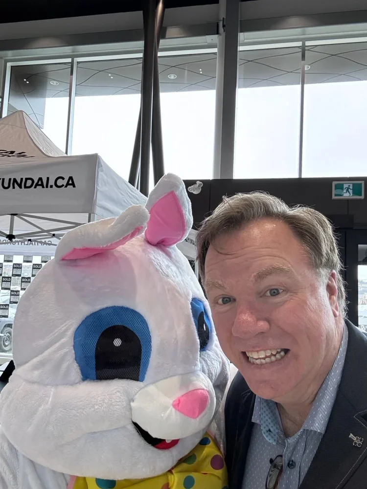 A man taking a selfie with a person dressed in an Easter Bunny costume with large blue eyes, pink nose, and a yellow bow tie with colorful polka dots, inside an airport terminal.