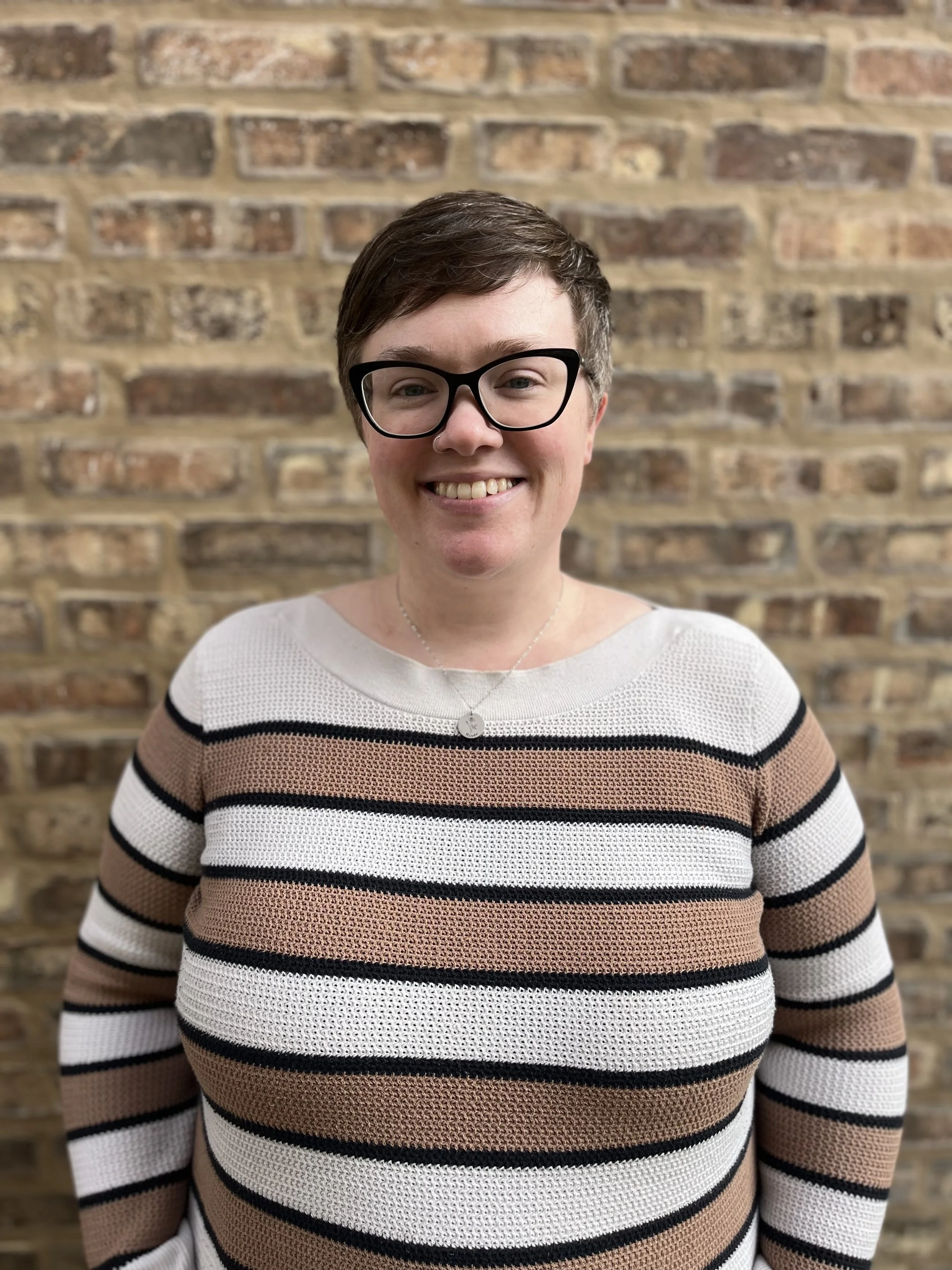 Photo of Dr. Ashley Molin wearing a brown and white sweater and glass while standing in front of a brick wall.